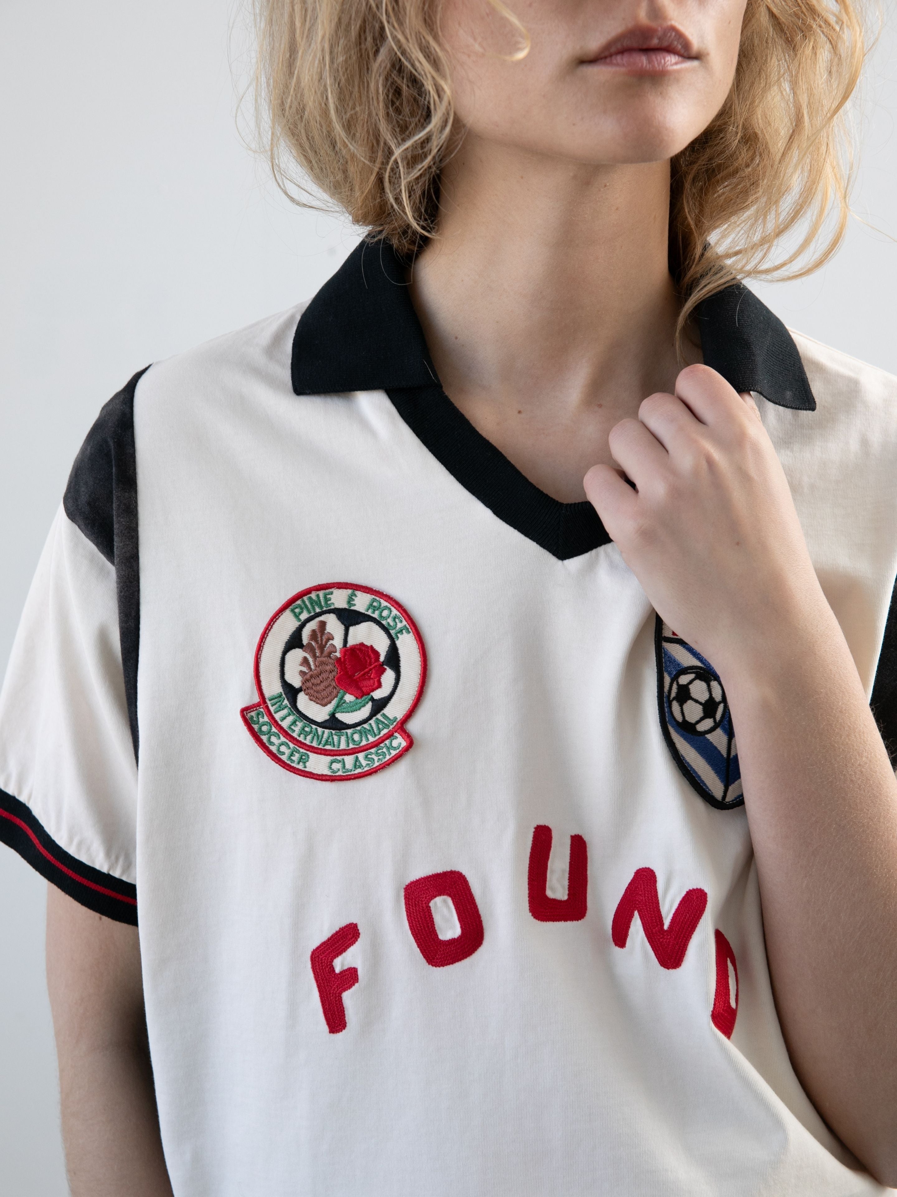 A blonde person wears the FOUND Union Soccer Jersey, a white unisex shirt with a black collar and sleeves, red "FOUND" lettering, and two embroidered chest patches.