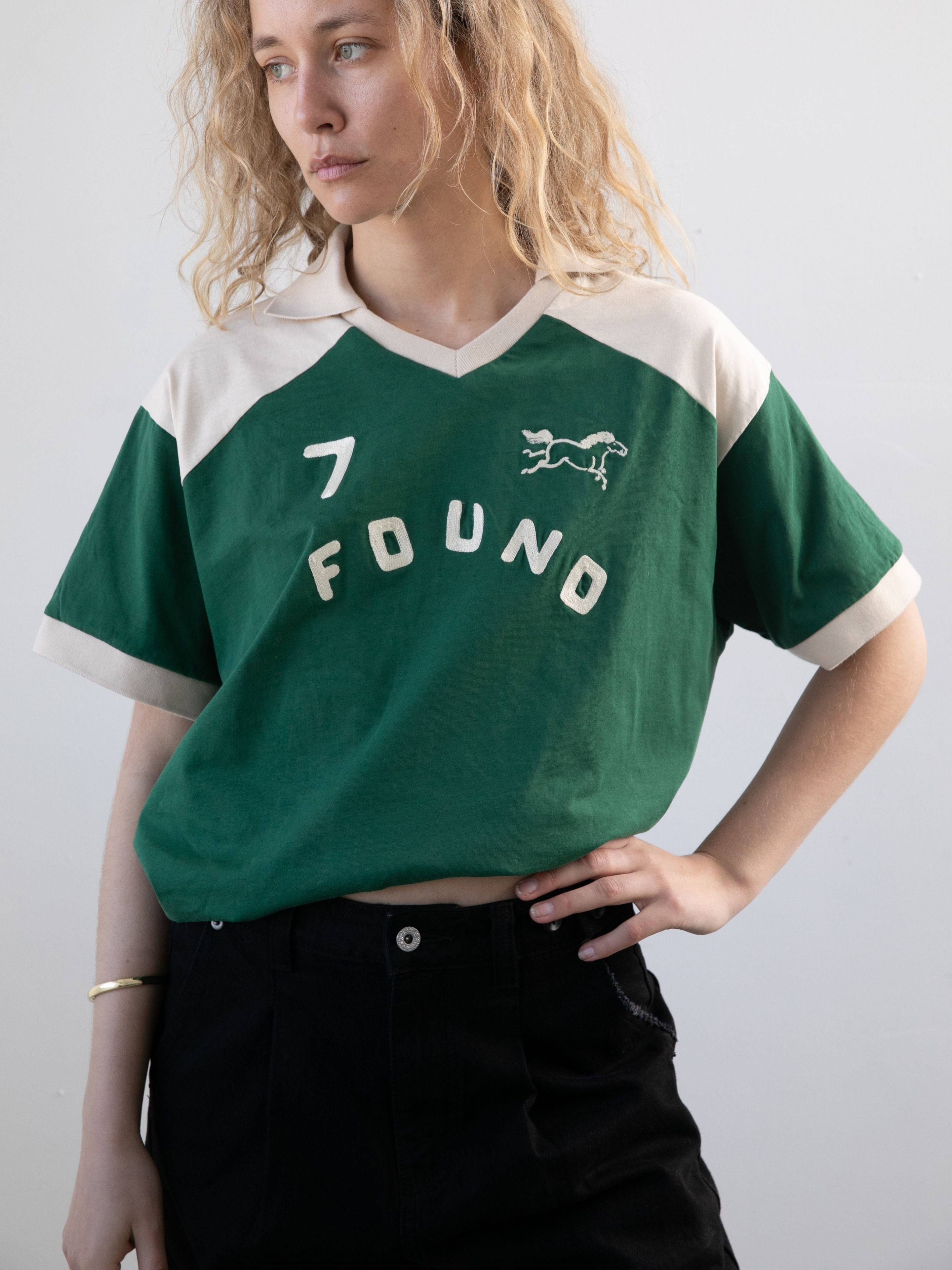 A person with wavy blonde hair wears the FOUND Goji Green Jersey featuring "7 FOUND" and a horse graphic, paired with unisex relaxed black pants, standing against a plain light background.