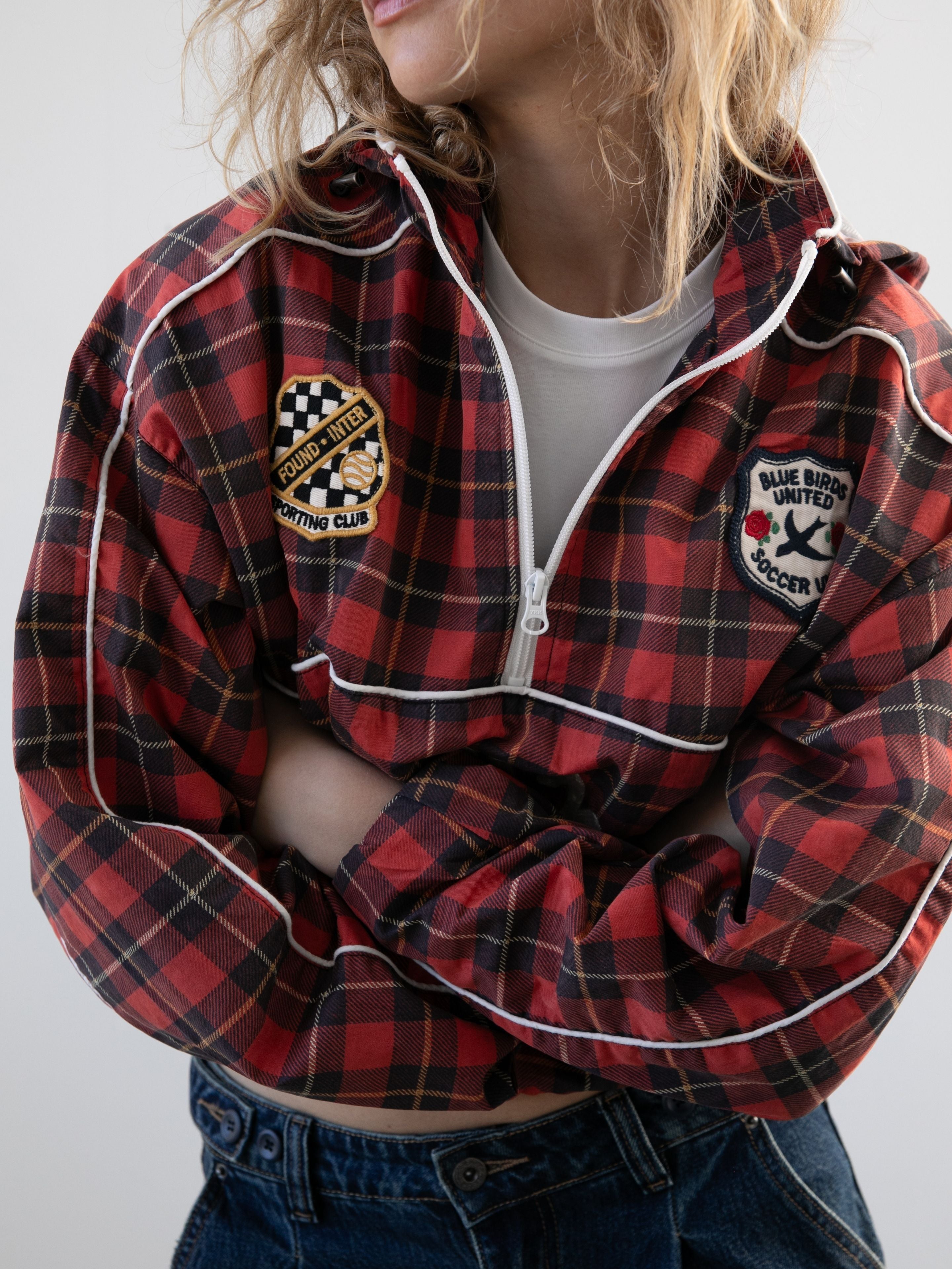 Model in the FOUND Red Plaid Track Jacket, layered over a white tee and blue jeans, stands with arms crossed. Face partially out of frame—channeling a classic vintage sport style.