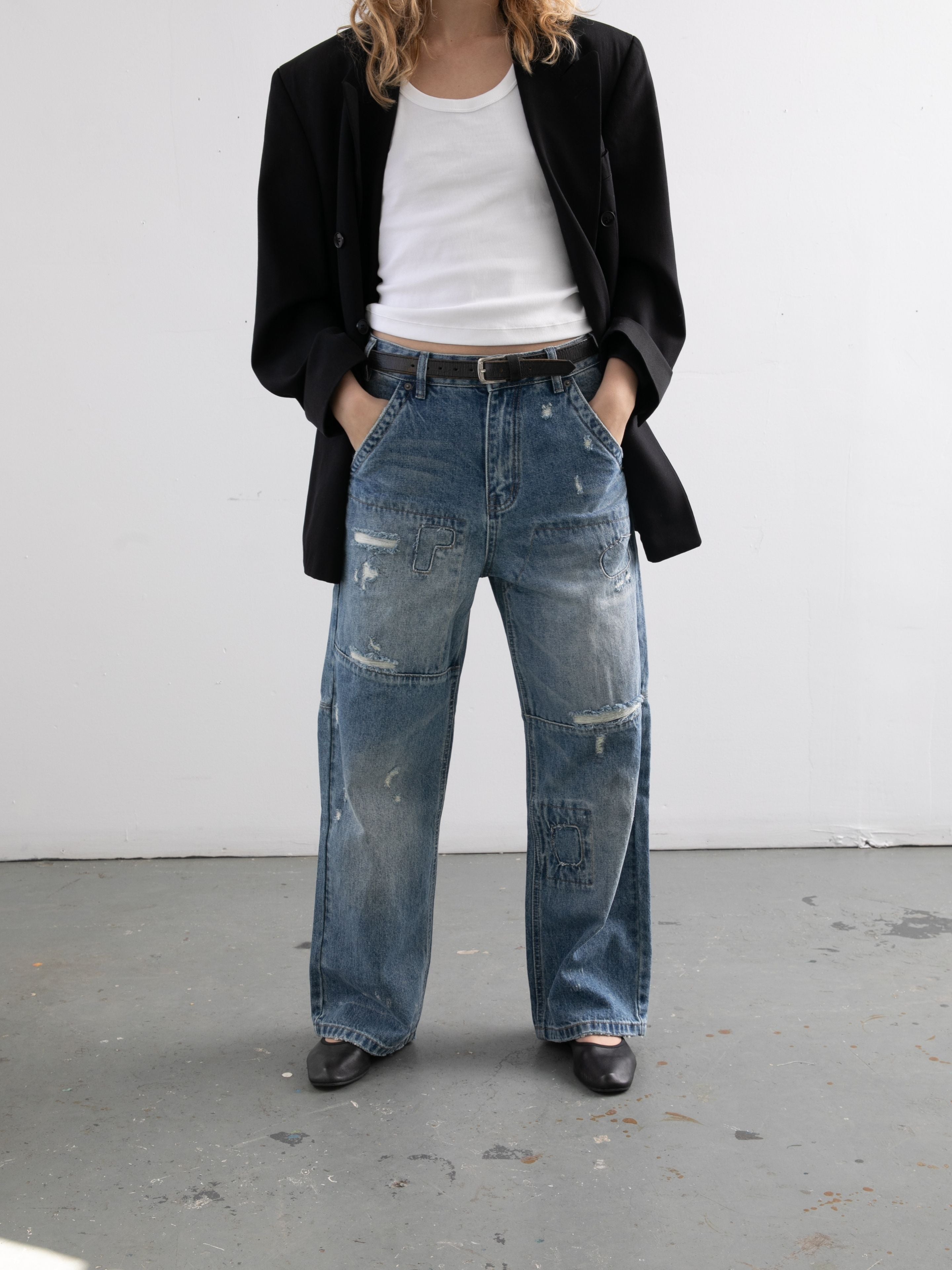 A person stands against a light wall with hands in pockets, wearing FOUND’s Painter Repaired Jeans, a black blazer, a white tank top, and black shoes.