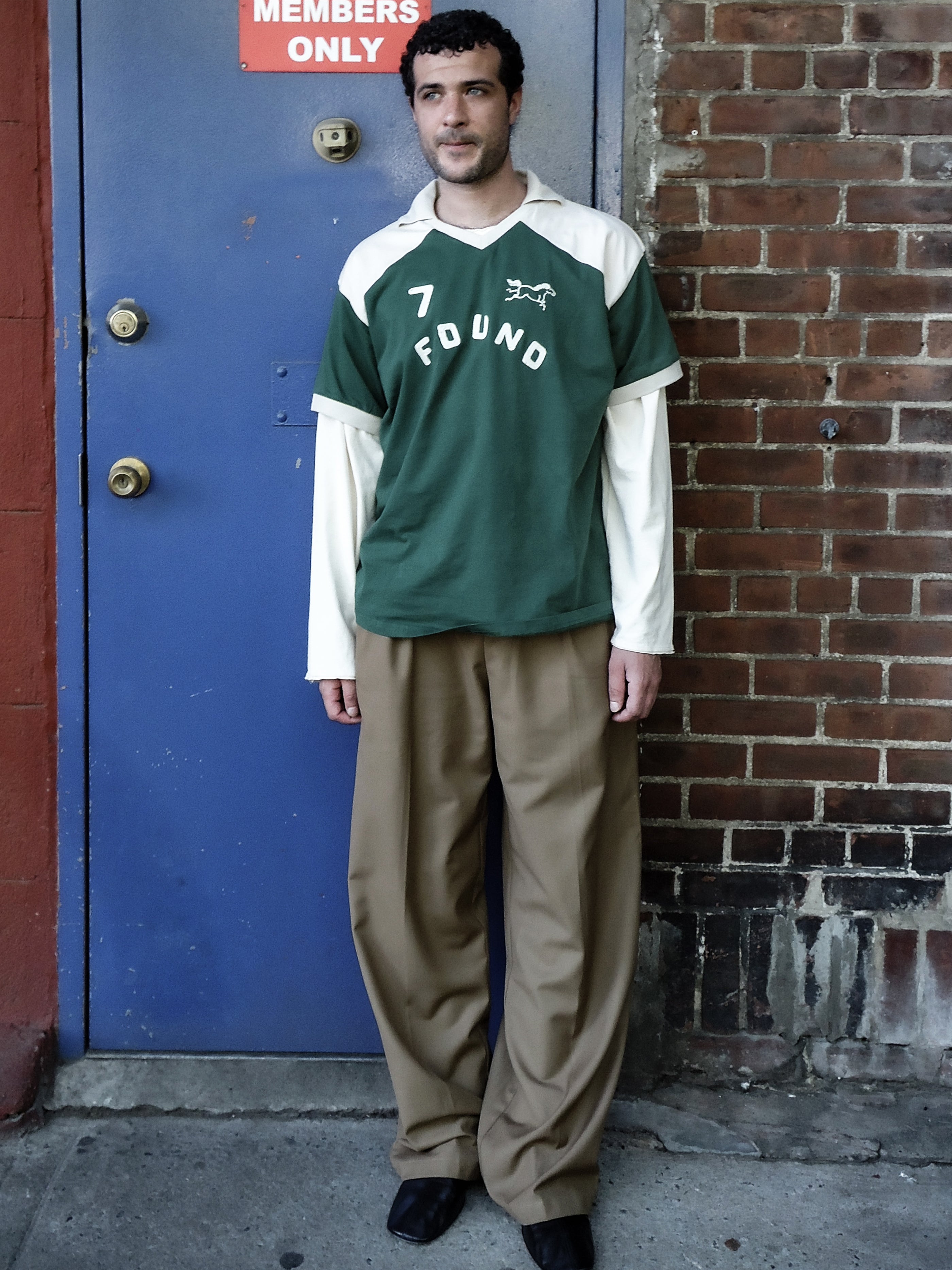 A man stands on a sidewalk in front of a blue door, wearing FOUND’s Goji Green Jersey over a long-sleeved top and loose brown pants, channeling the relaxed style of vintage athletic uniforms.