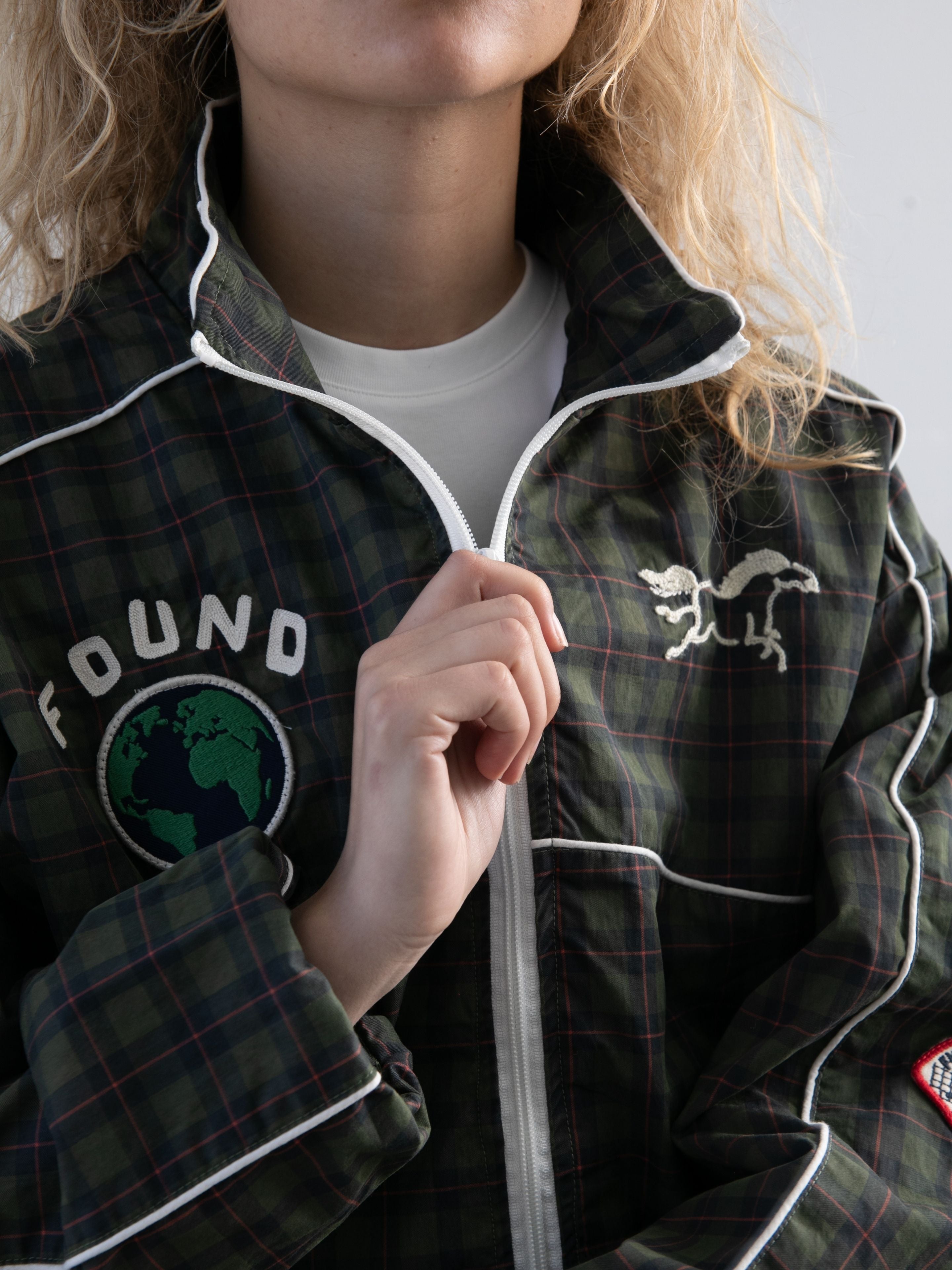 A person zips up the FOUND Forest Plaid Track Jacket, featuring white piping, a globe patch, the "FOUND" logo, and a white animal patch.