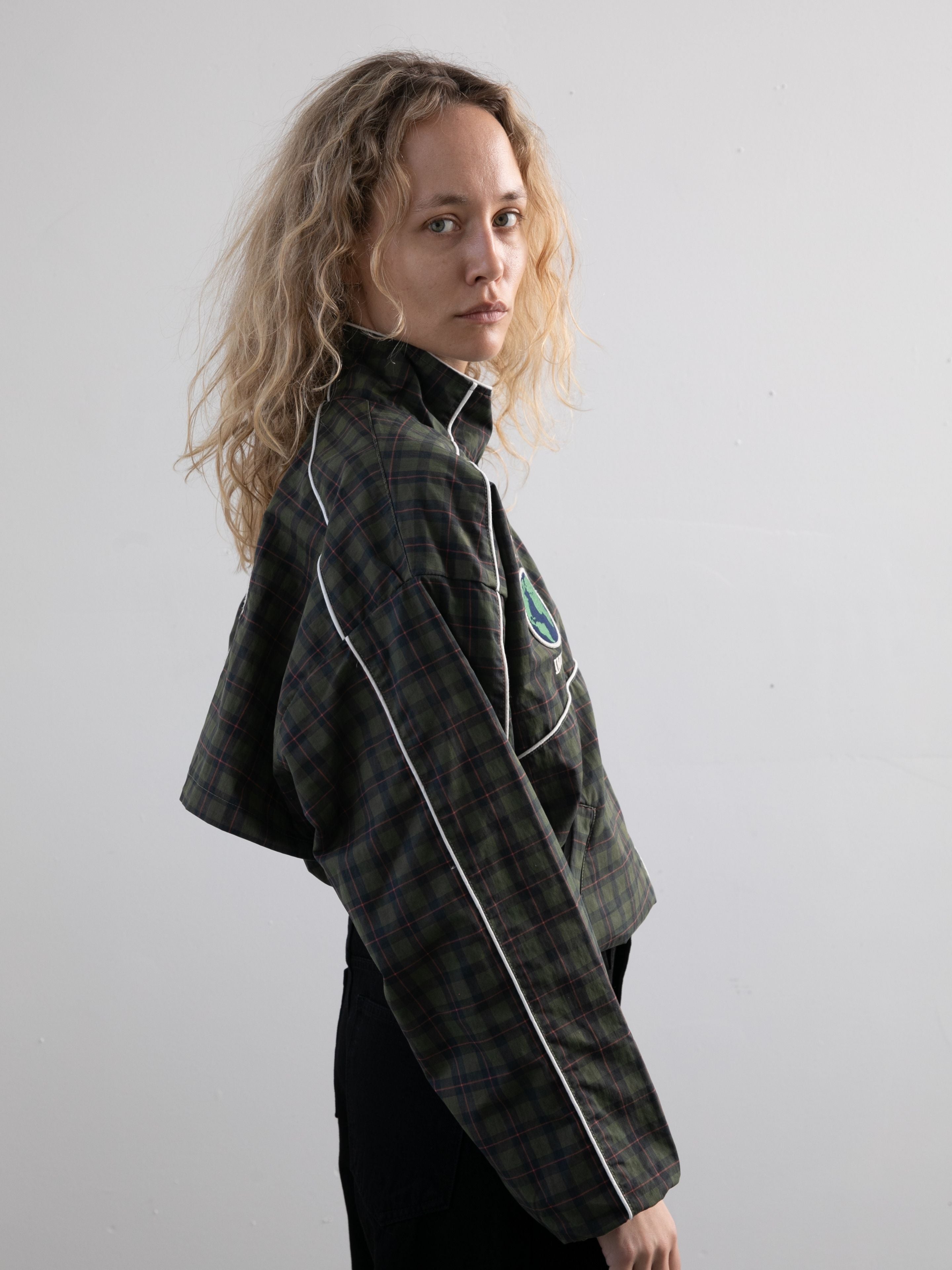 A person with wavy blonde hair wears the FOUND Forest Plaid Track Jacket, standing sideways and looking at the camera against a plain background.
