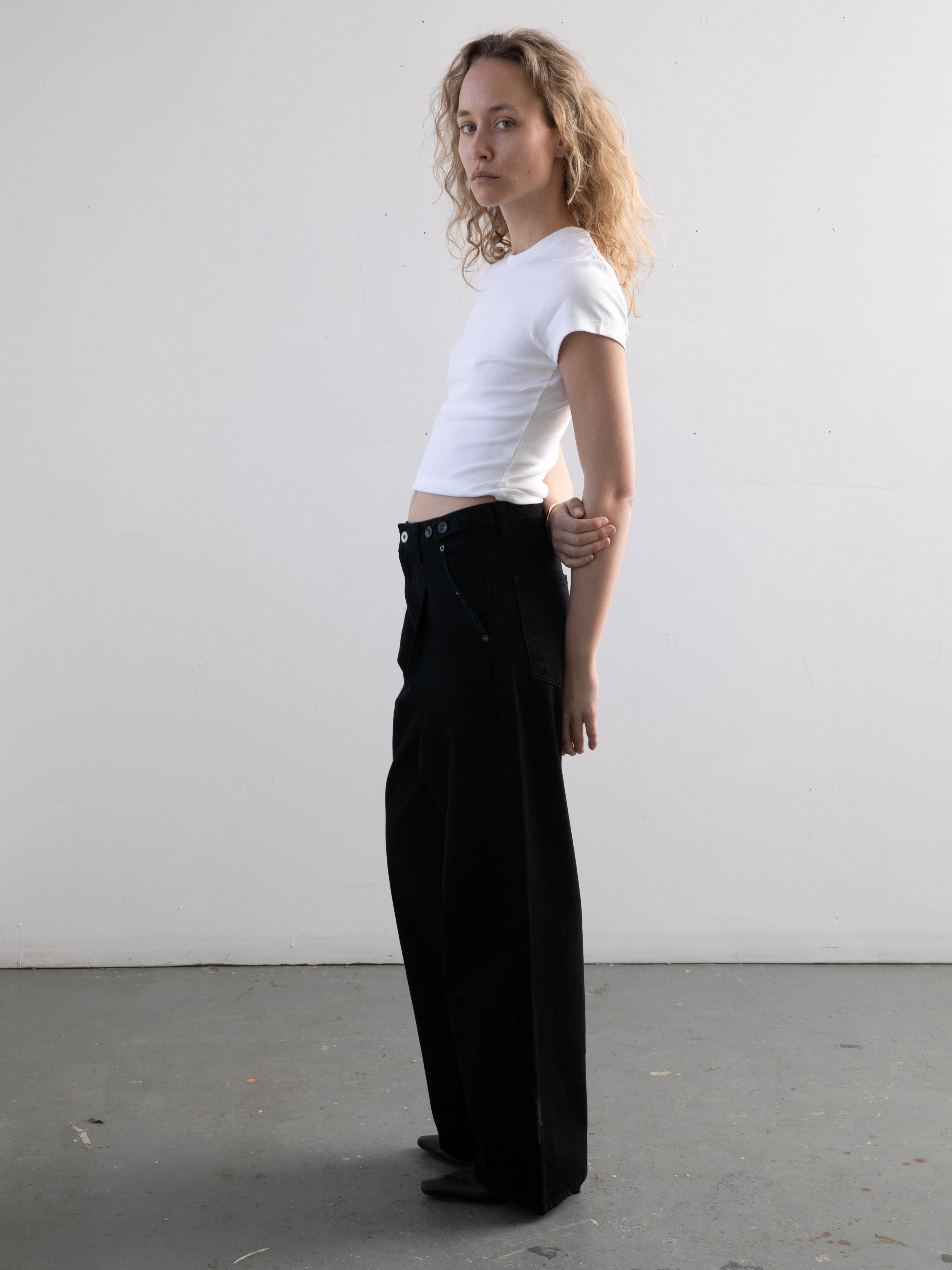 Person stands sideways by a plain white wall, wearing a fitted white t-shirt and Found Pleated Relaxed Fit Jeans with a loose, vintage denim style. Curly hair frames their neutral expression.