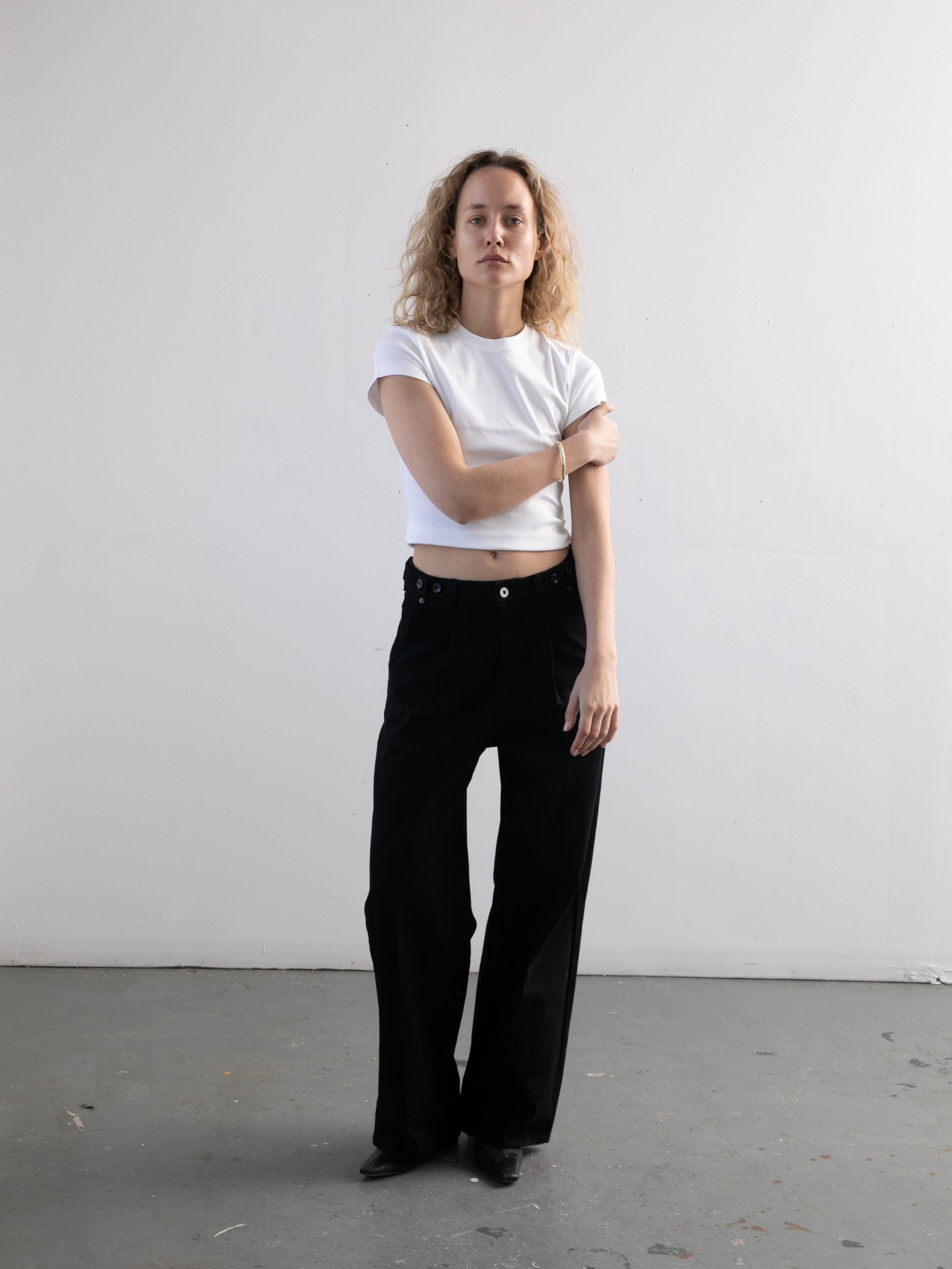 A person with curly hair stands against a plain white wall, wearing a white short-sleeve shirt and Found Pleated Relaxed Fit Jeans for a casual, timeless look.