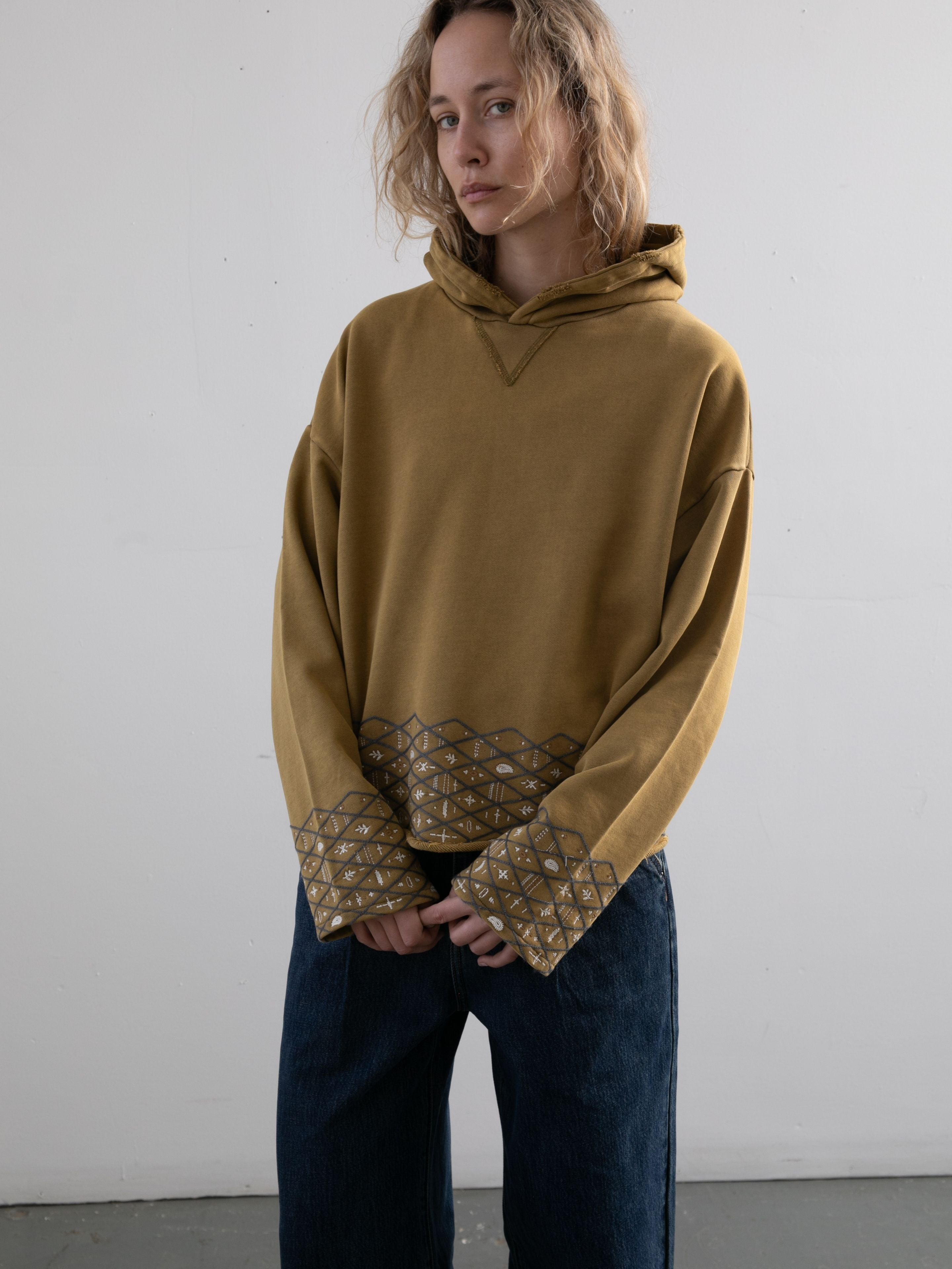 A person with shoulder-length blonde hair wears the FOUND Embroidered Border Hoodie—mustard yellow with patterned cuffs and hem—and blue jeans, standing against a plain light-colored wall.