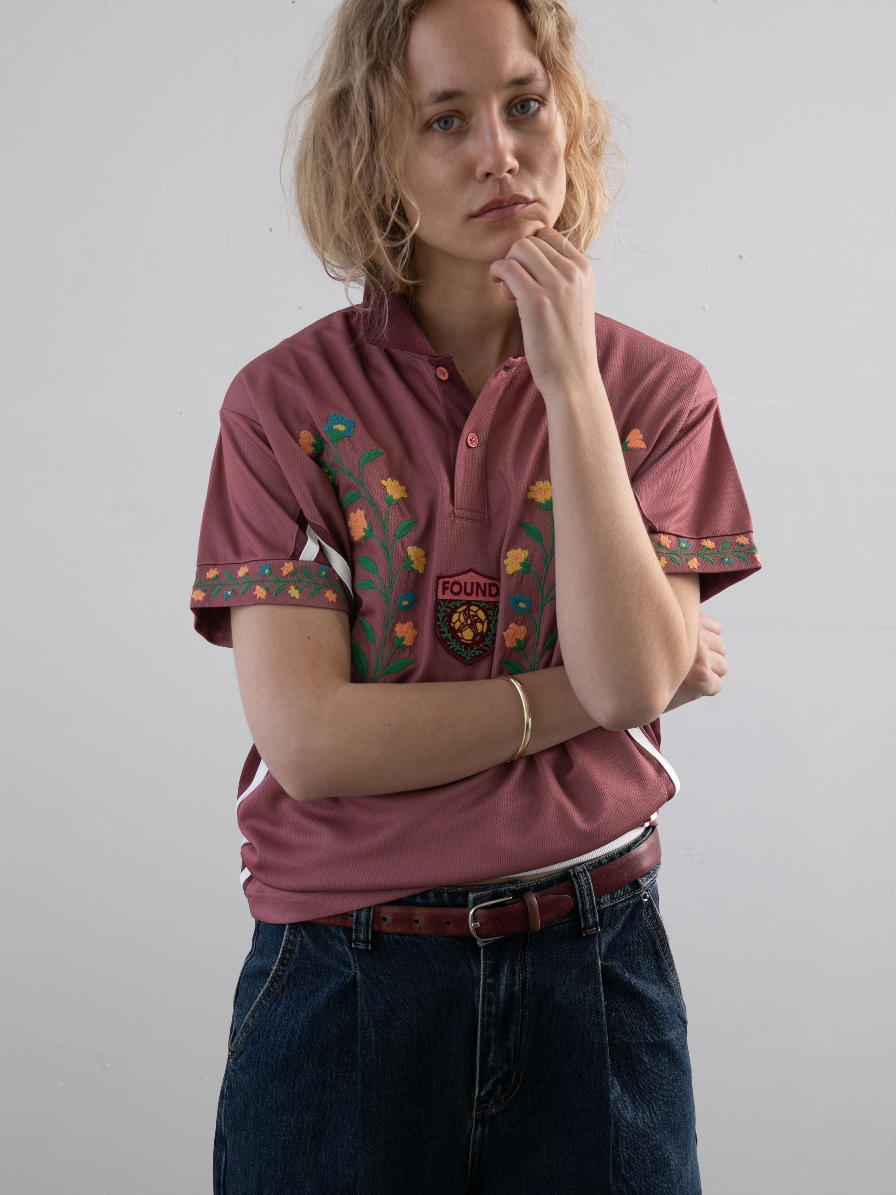 A person with short blond hair wears the FOUND Floral Polo Jersey and blue jeans, standing against a plain white wall with one hand touching their face.