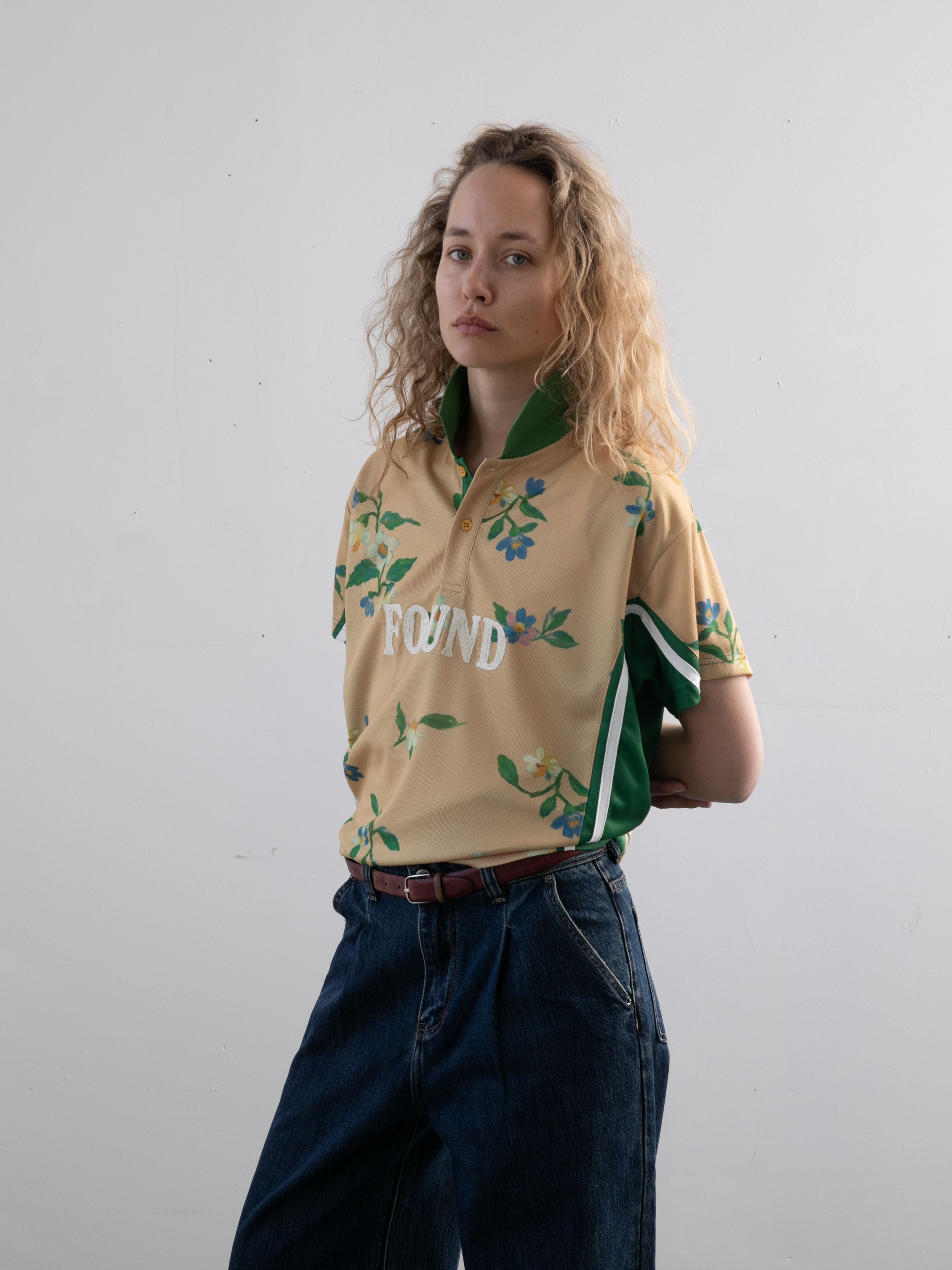A person with wavy blonde hair wears a beige FOUND Floral Polo Jersey and blue jeans, standing against a plain white background.