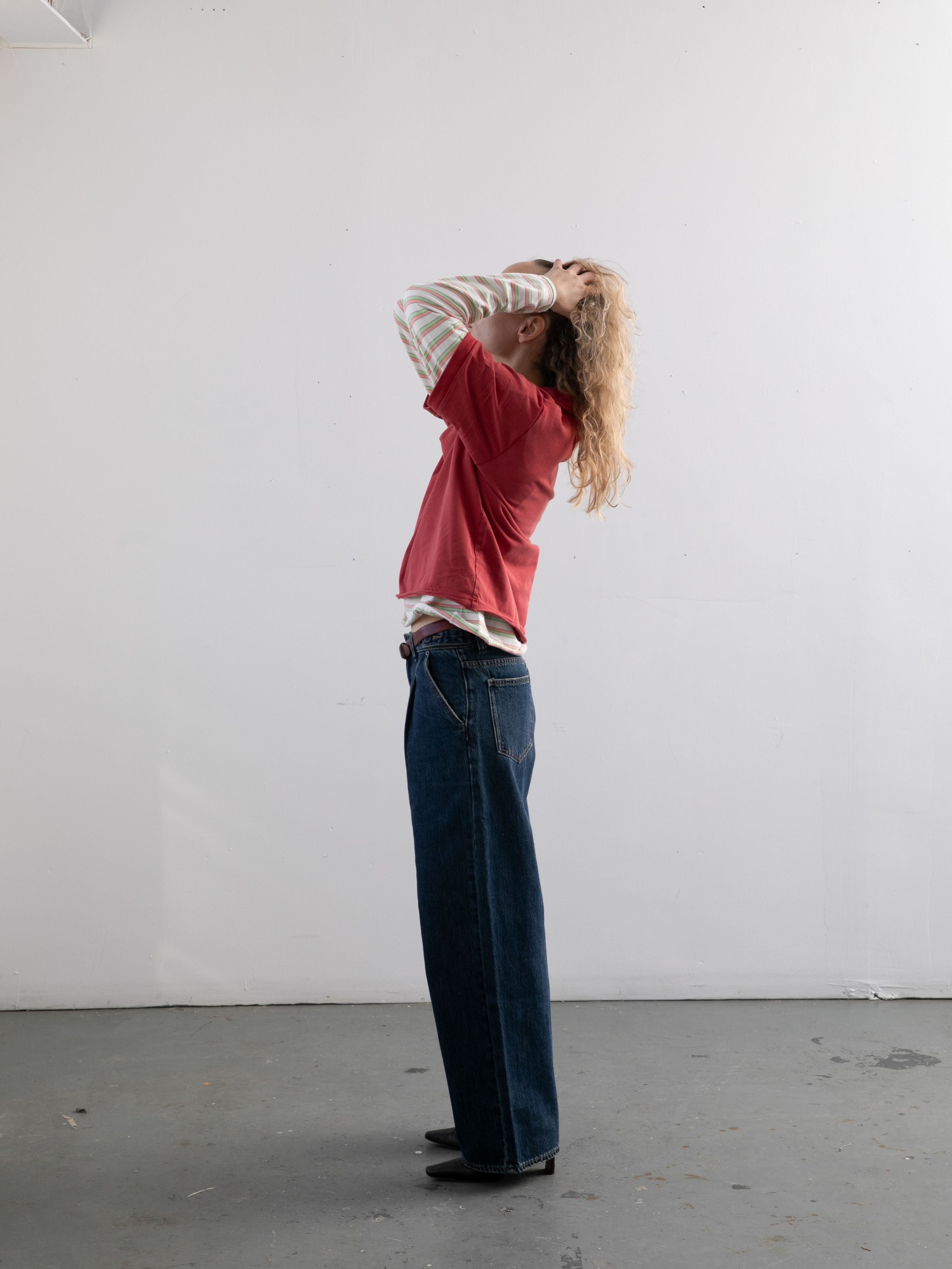 A person stands sideways against a white wall, hands on head, wearing blue jeans, black shoes, and a FOUND Double Layer Stripe Polo over a red T-shirt—an effortless unisex sportswear look.