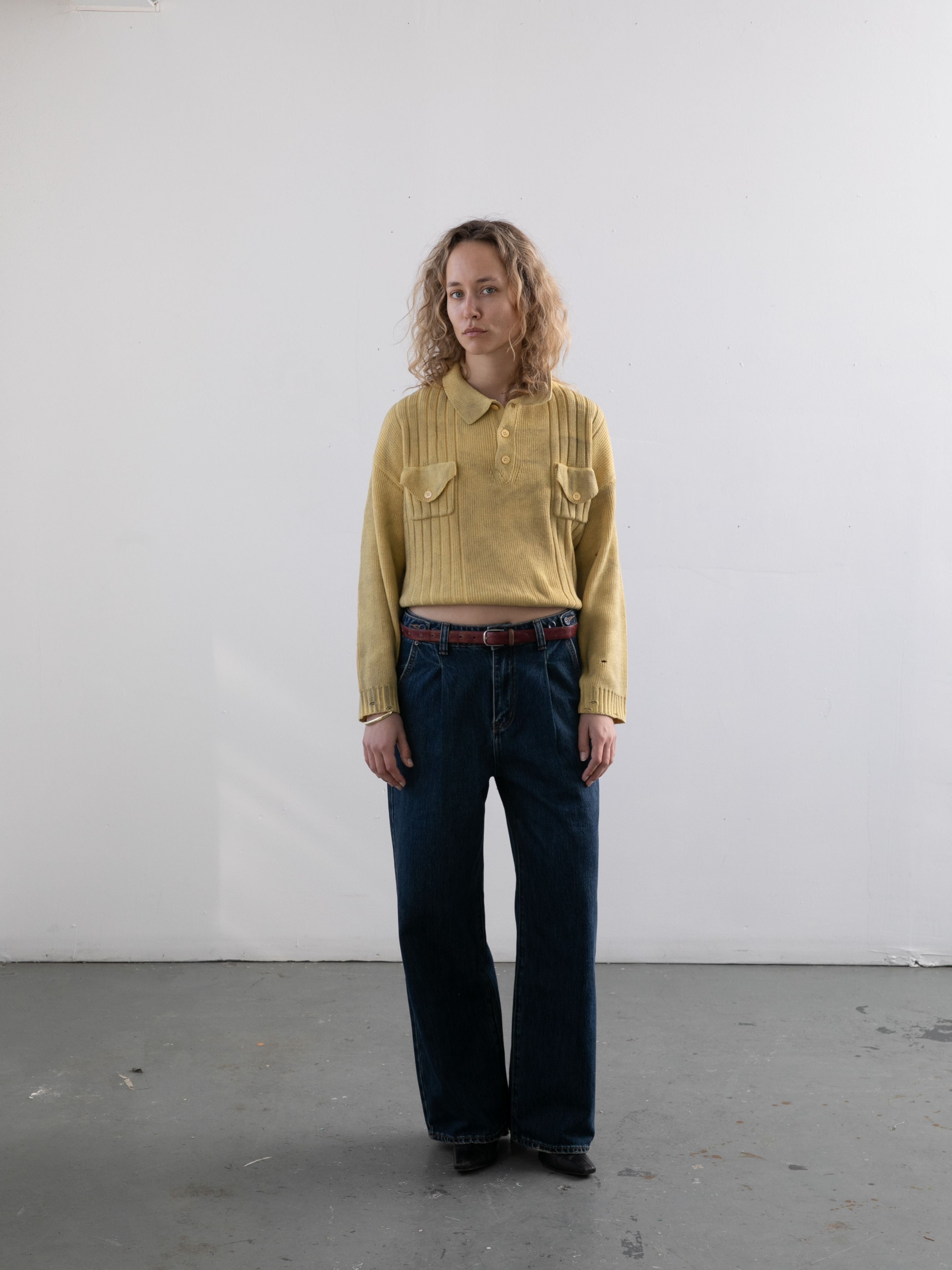 A person with curly hair stands indoors against a plain white wall, wearing the FOUND Distressed Pullover Knit and blue jeans—versatile knitwear for any season.