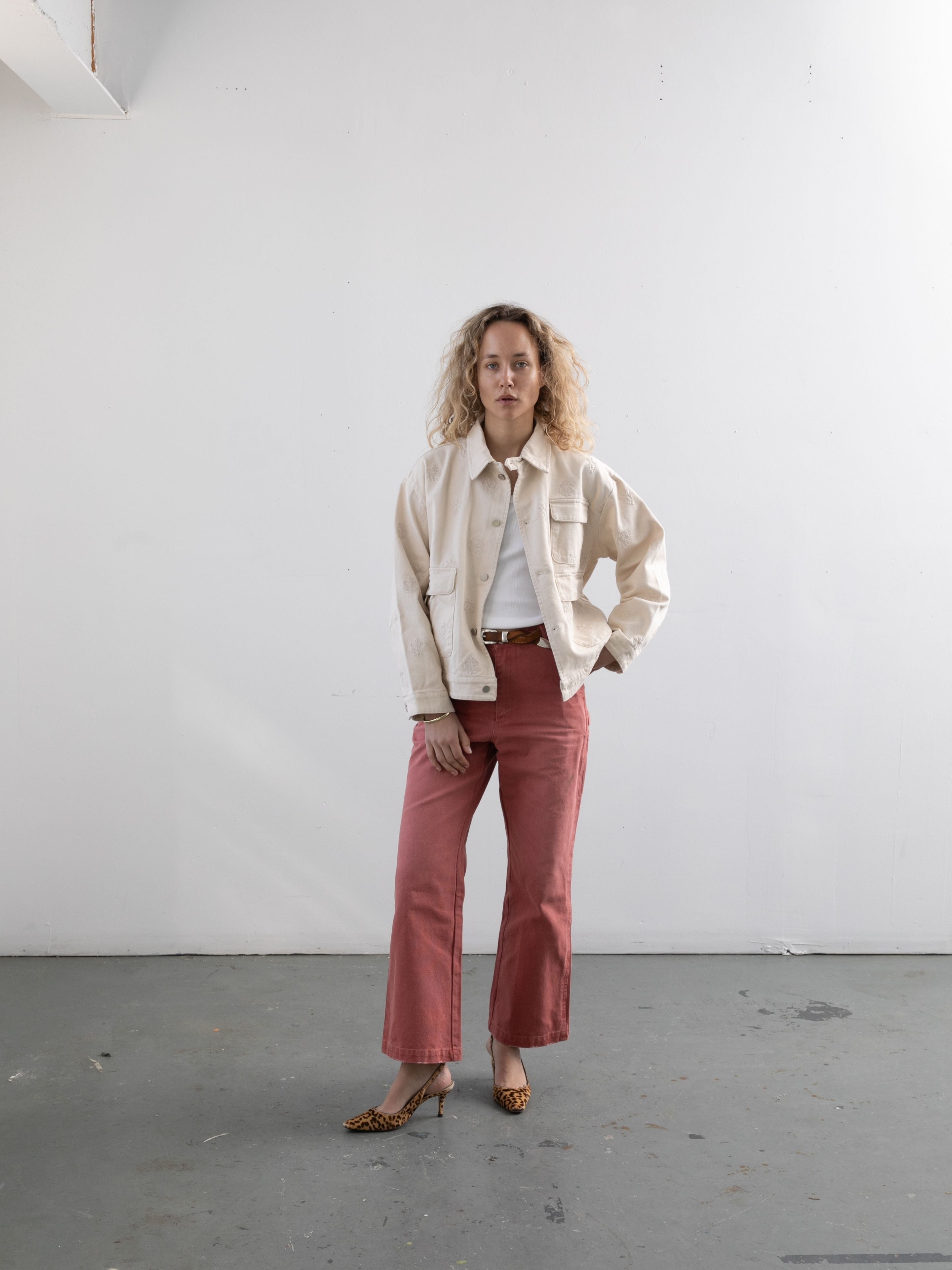 A woman with curly hair stands in a minimalist room, wearing Found's Embroidered Motif Jacket over a white shirt, paired with pink pants and leopard print heels.