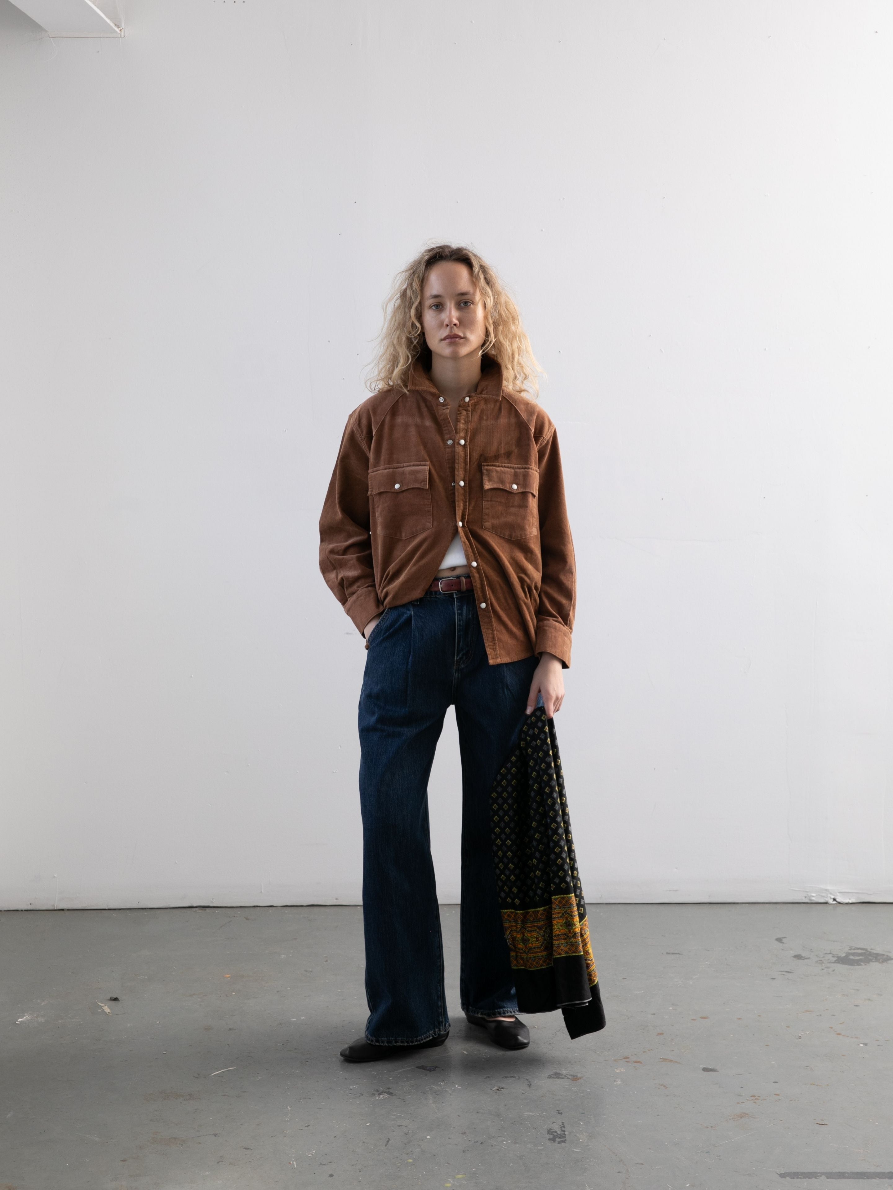 A person with curly blond hair stands indoors against a white wall, wearing the FOUND Western Corduroy Shirt and blue jeans, holding a black and yellow patterned fabric.