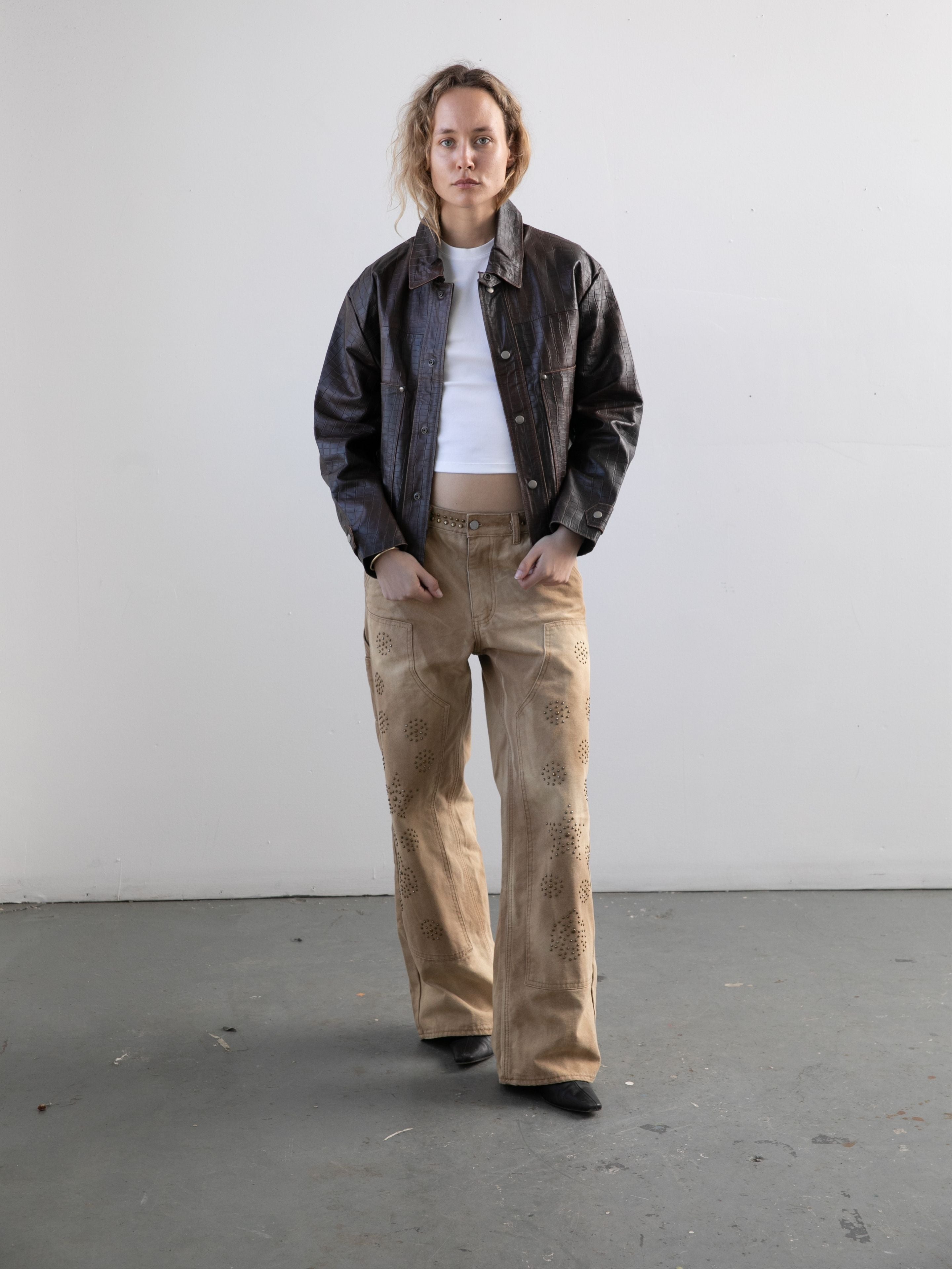 A person stands in a plain room wearing the FOUND Distressed Croc Embossed Leather Work Jacket, a white turtleneck, tan patterned pants, and black shoes, facing the camera with a neutral expression.