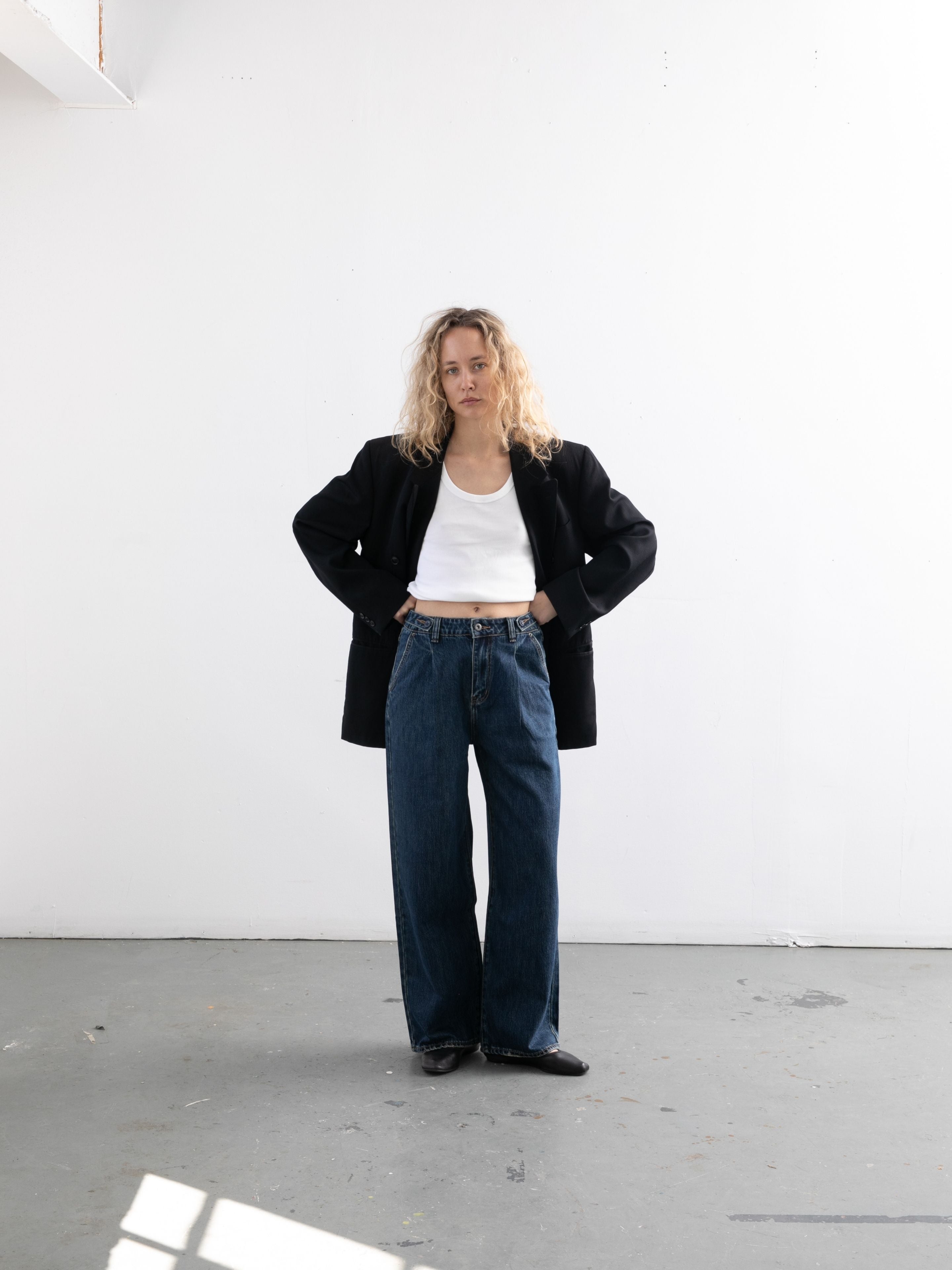 A woman with curly blonde hair stands indoors against a white wall, wearing a black blazer, white tank top, blue Found Pleated Relaxed Fit Jeans, and black shoes.