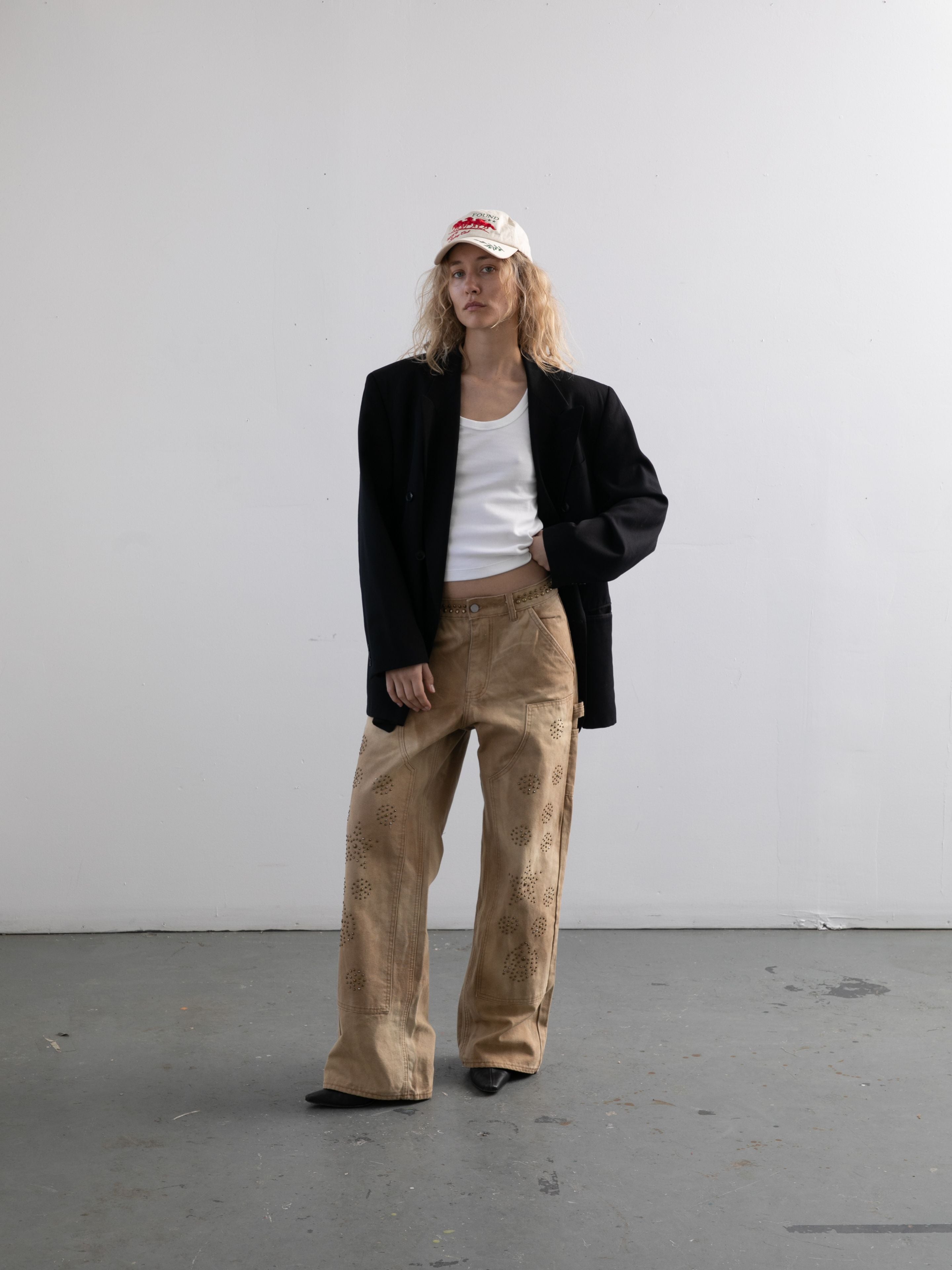 A person stands against a plain white wall wearing a white baseball cap, black blazer, white shirt, and FOUND Canvas Studded Work Pants.