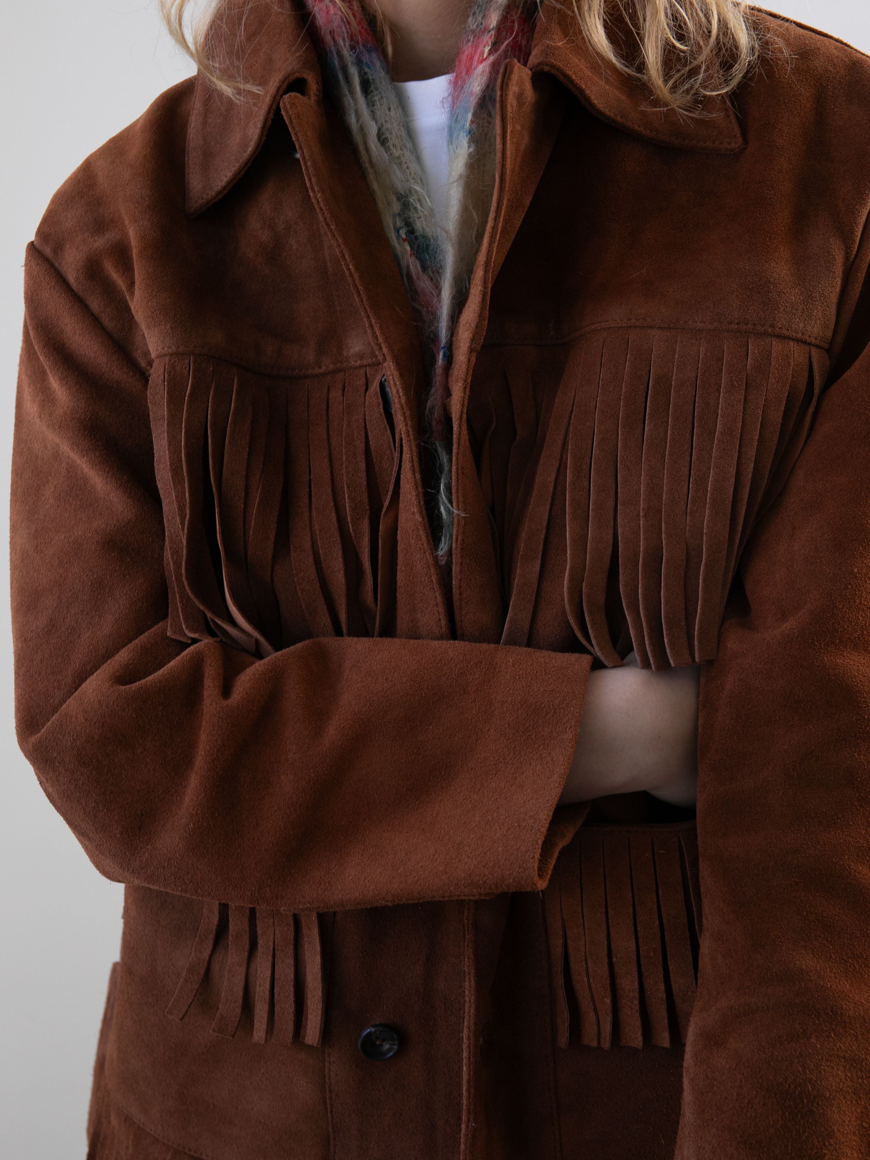 A person wearing the FOUND Western Suede Fringe Jacket with fringe details and a colorful scarf, arms crossed.
