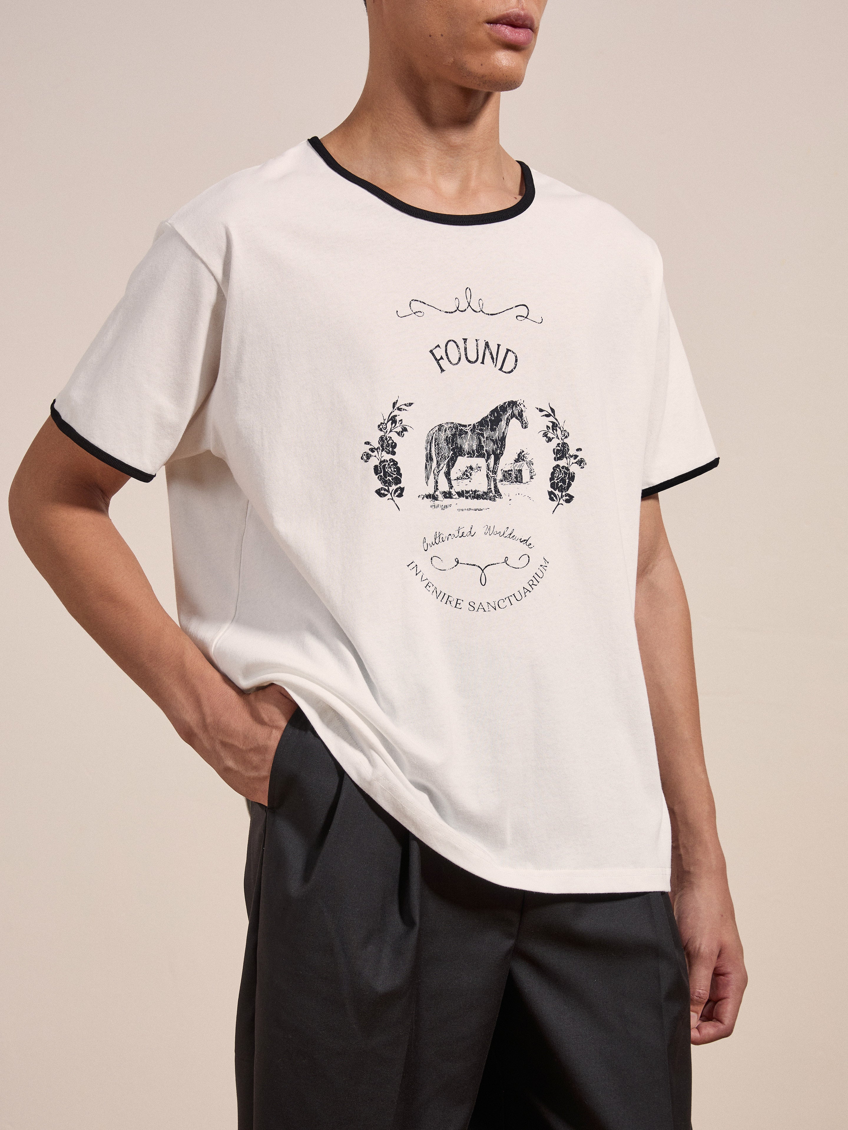 A person is wearing a Horse Crest Ringer Tee by FOUND, which is a white T-shirt with black trim featuring a graphic of a tiger and the text "FOUND" and "FUNDING SANCTUARIES." They have one hand in their pocket and are wearing dark pants.