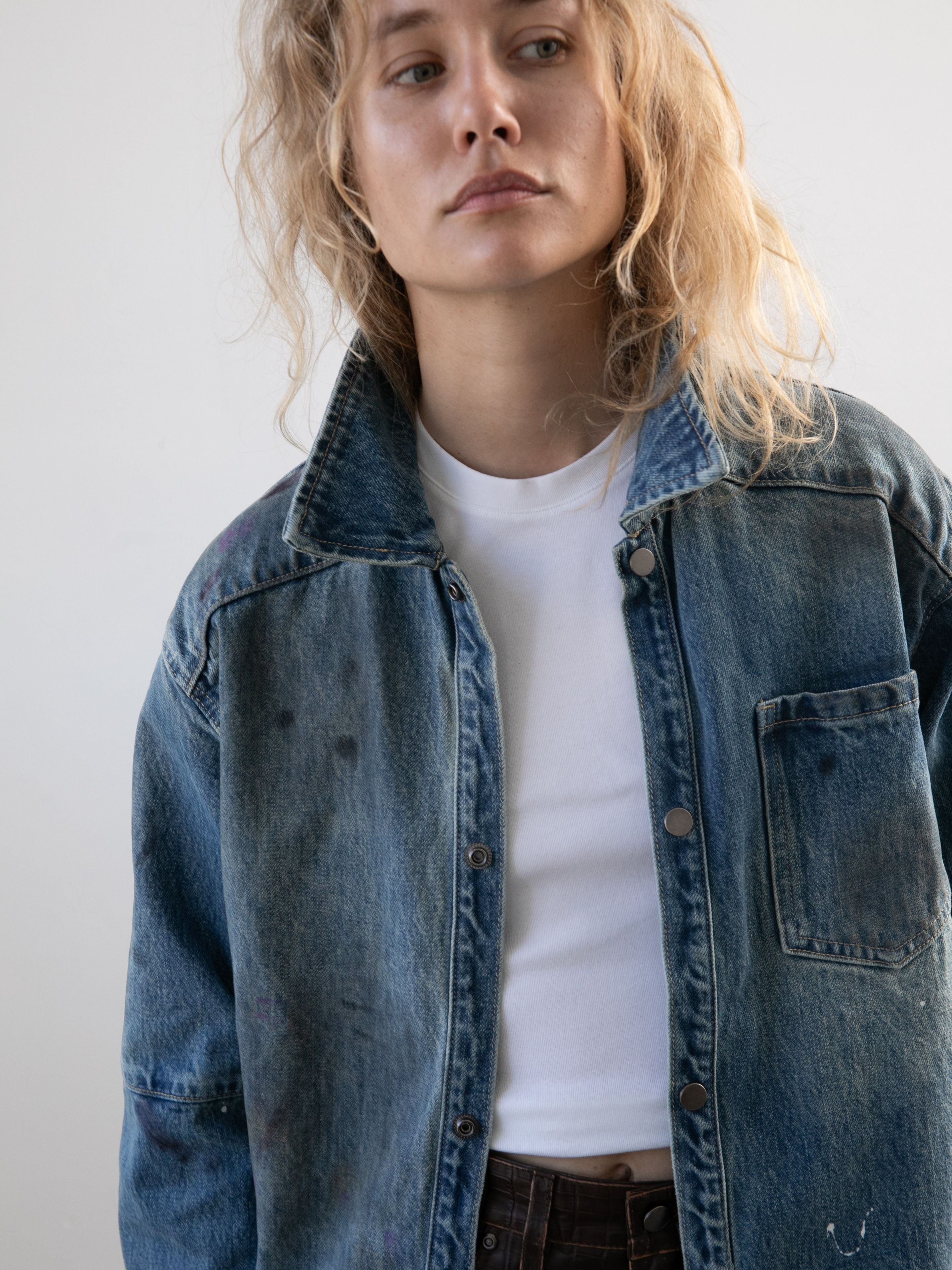 A person with wavy blonde hair wears the FOUND Western Denim Painter Shirt over a white tee, standing against a plain light background and glancing to the side.