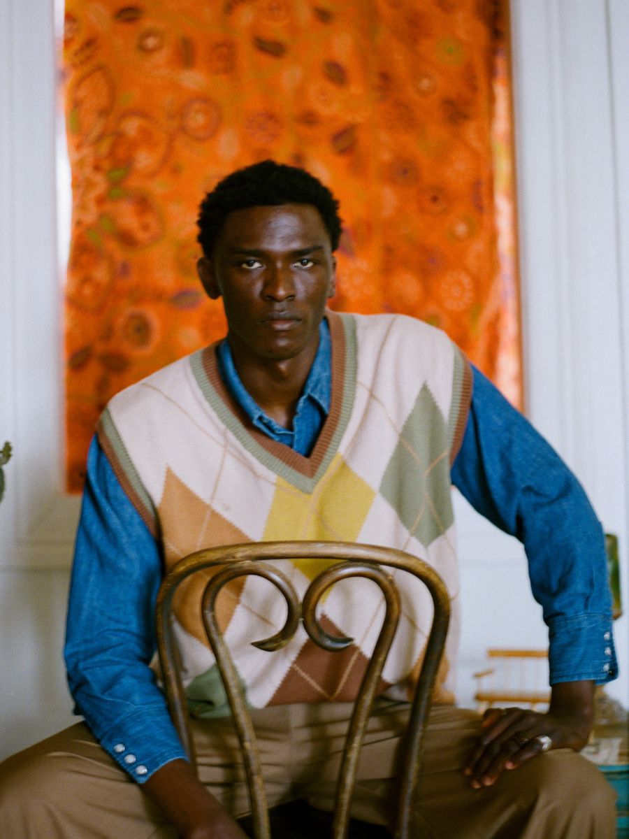 A person sits on a wooden chair wearing a blue shirt and FOUND's Argyle Knit Vest, surrounded by an orange patterned curtain that enhances the collegiate vibe.