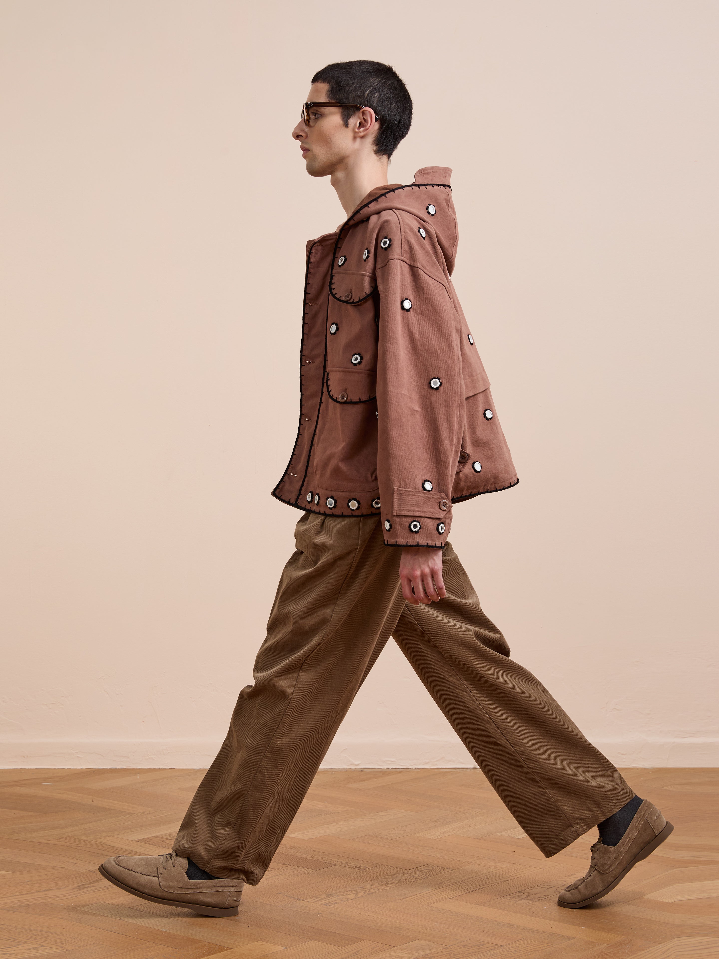 A person in glasses walks indoors, wearing the FOUND Hand Embroidered Hooded Mirror Anorak with circular mirror-inspired details, loose brown pants, and brown shoes on a plain background with a wooden floor.