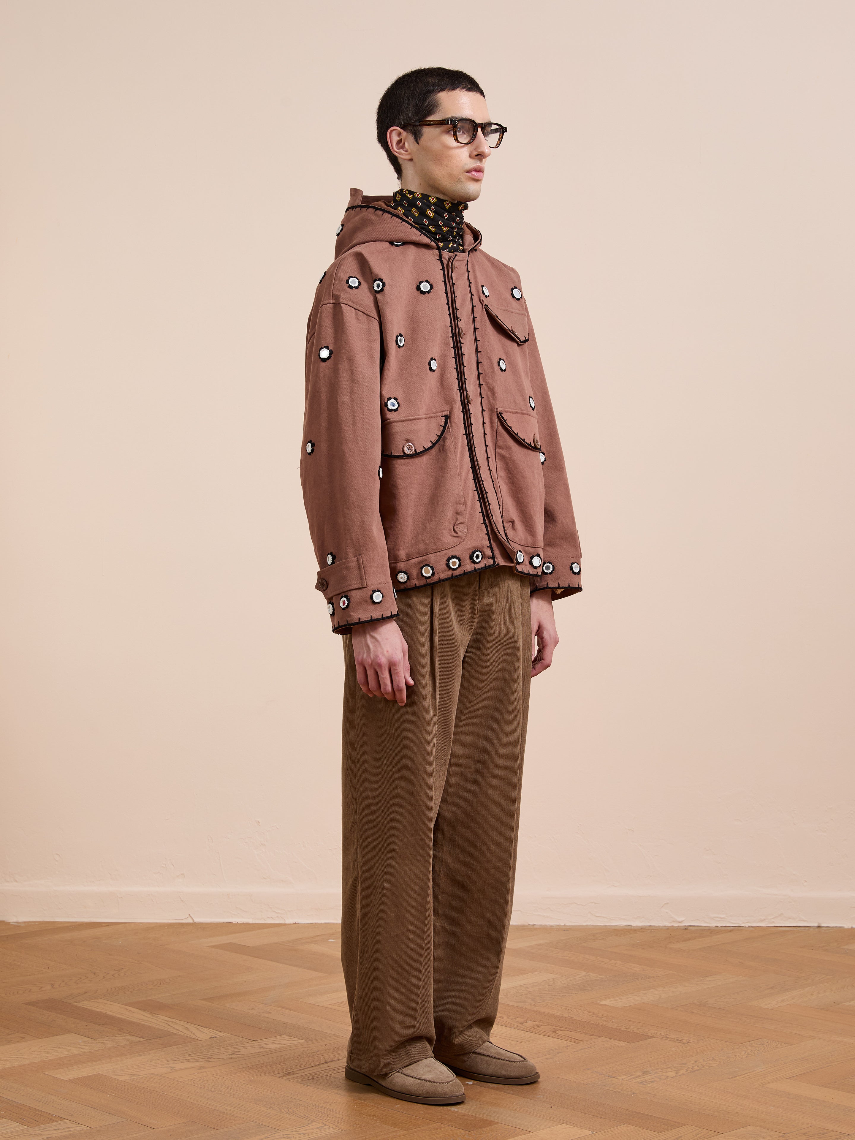 Indoors against a plain background, a person wears FOUND's Hand Embroidered Hooded Mirror Anorak with glasses, a patterned turtleneck, brown wide-leg pants, and beige shoes.