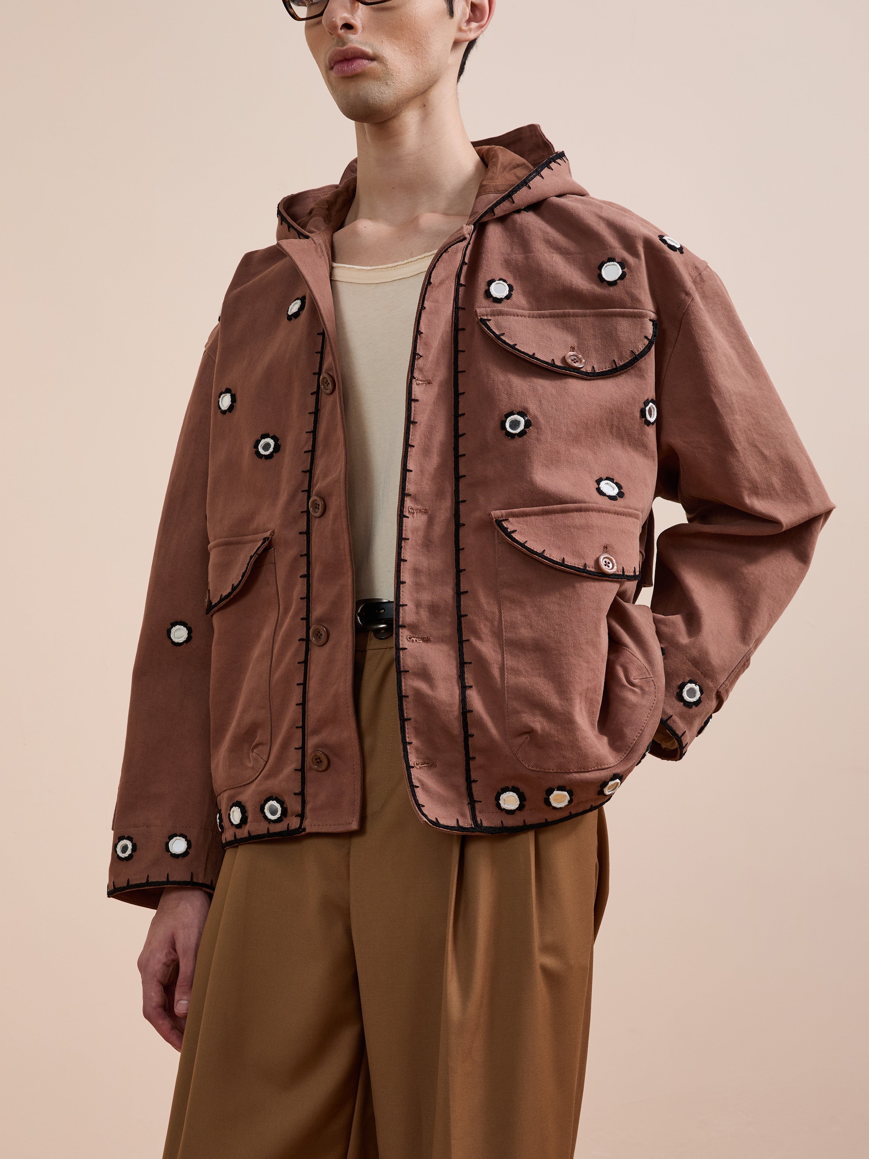 A person wears the FOUND Hand Embroidered Hooded Mirror Anorak, featuring black trim and white mirror-inspired accents, paired with a beige shirt and tan trousers, standing against a neutral background.