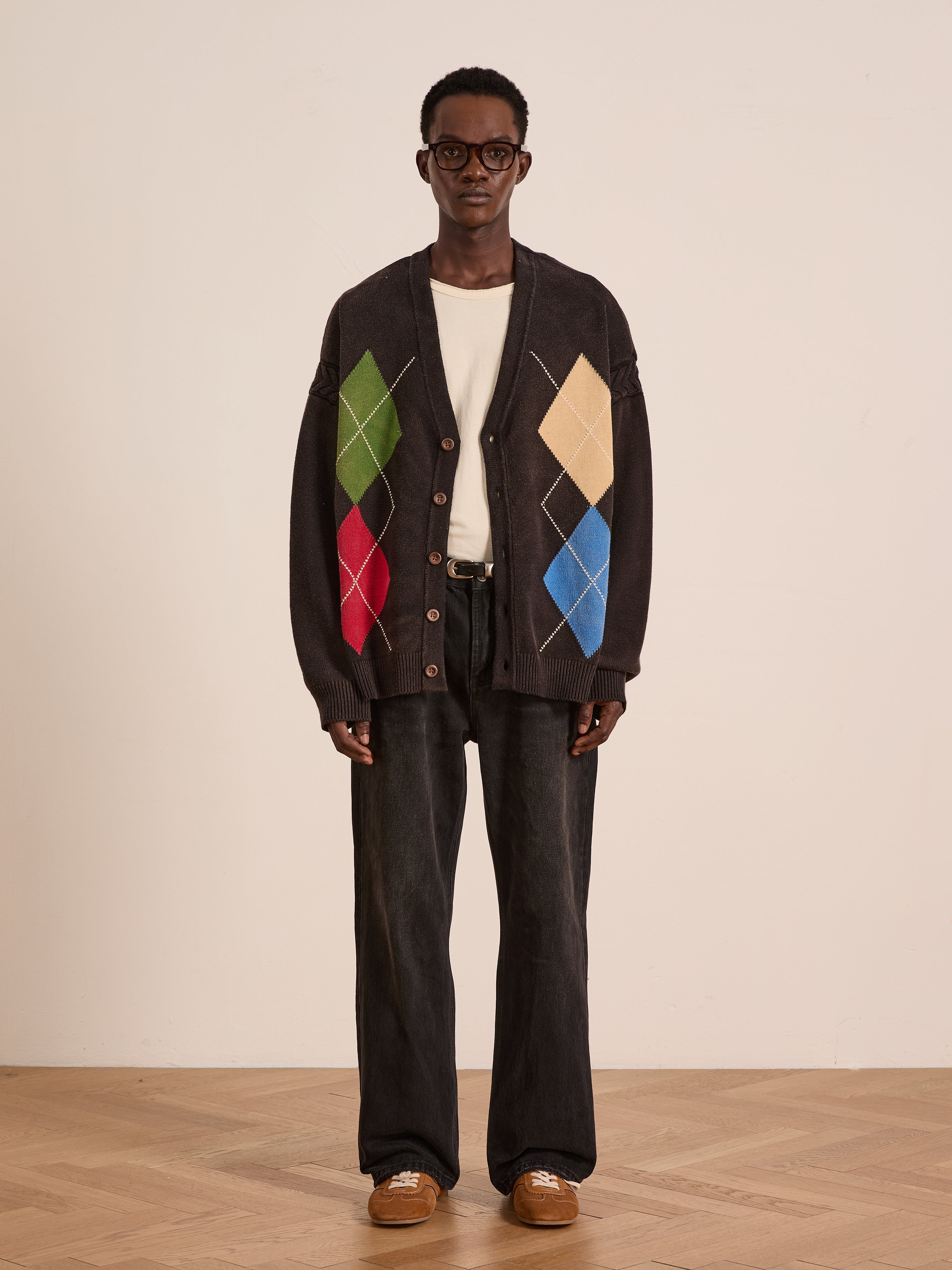 Indoors, a person wears glasses, a white shirt, wide-leg dark jeans, brown shoes, and the FOUND Argyle Knit Cardigan with colorful diamond patterns.