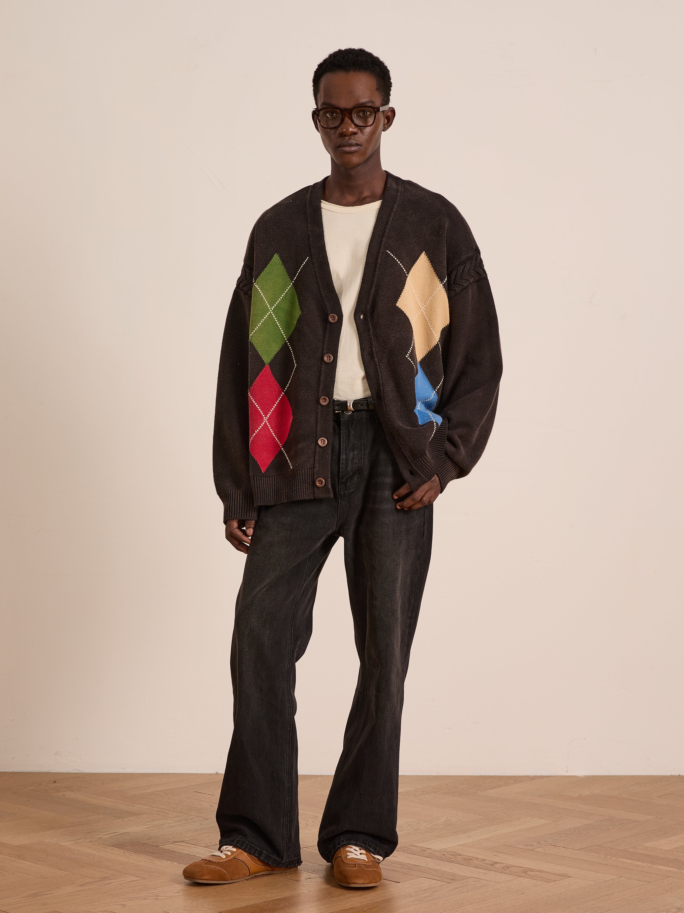 Indoors, a person wears glasses, a white shirt, black jeans, brown shoes, and the FOUND Argyle Knit Cardigan with colorful patterns, standing on a wooden floor against a plain light wall.