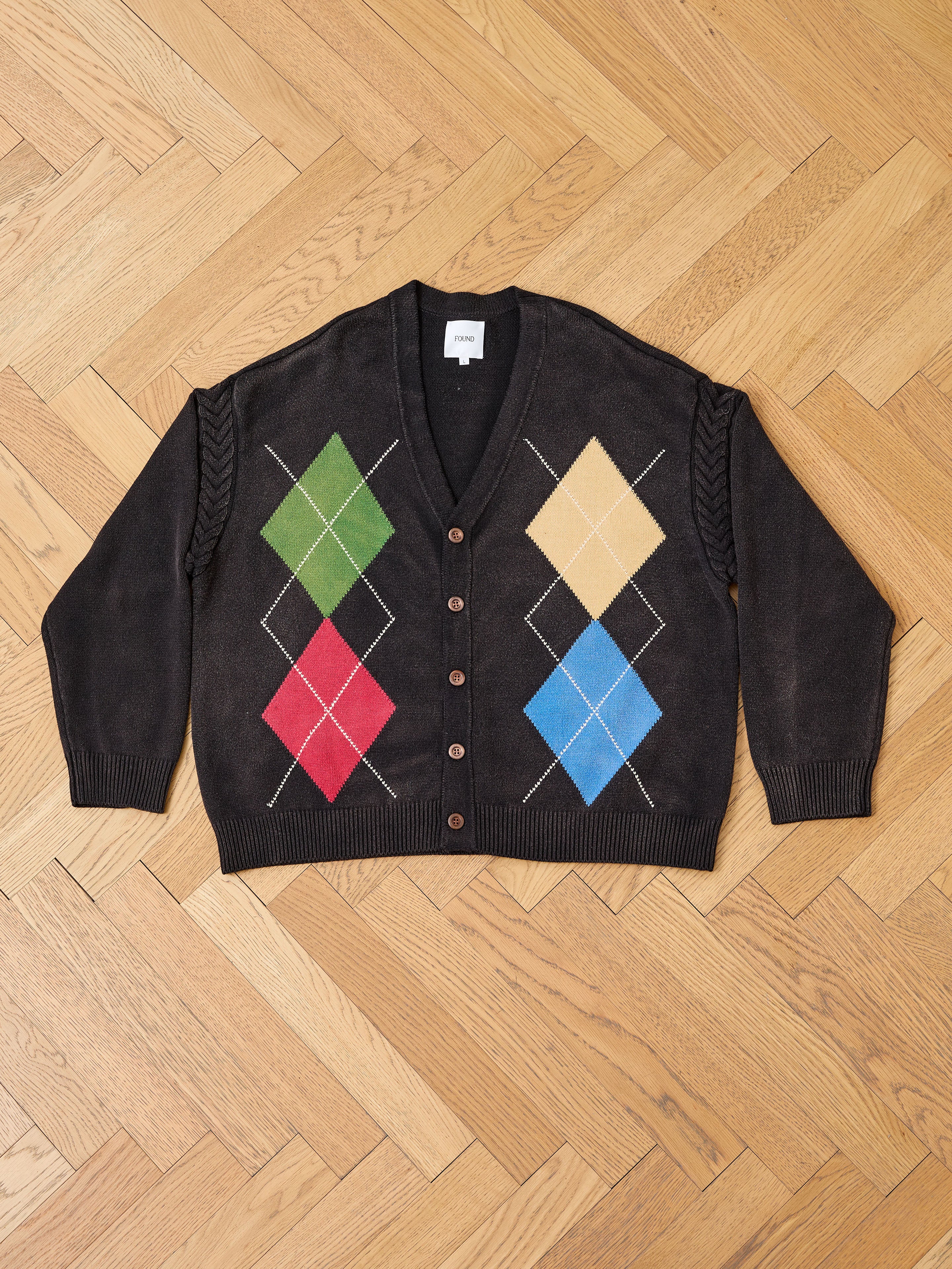 The FOUND Argyle Knit Cardigan features an oversized fit, front button closure, and a multicolored diamond pattern in black, shown laid flat on a wooden herringbone floor.