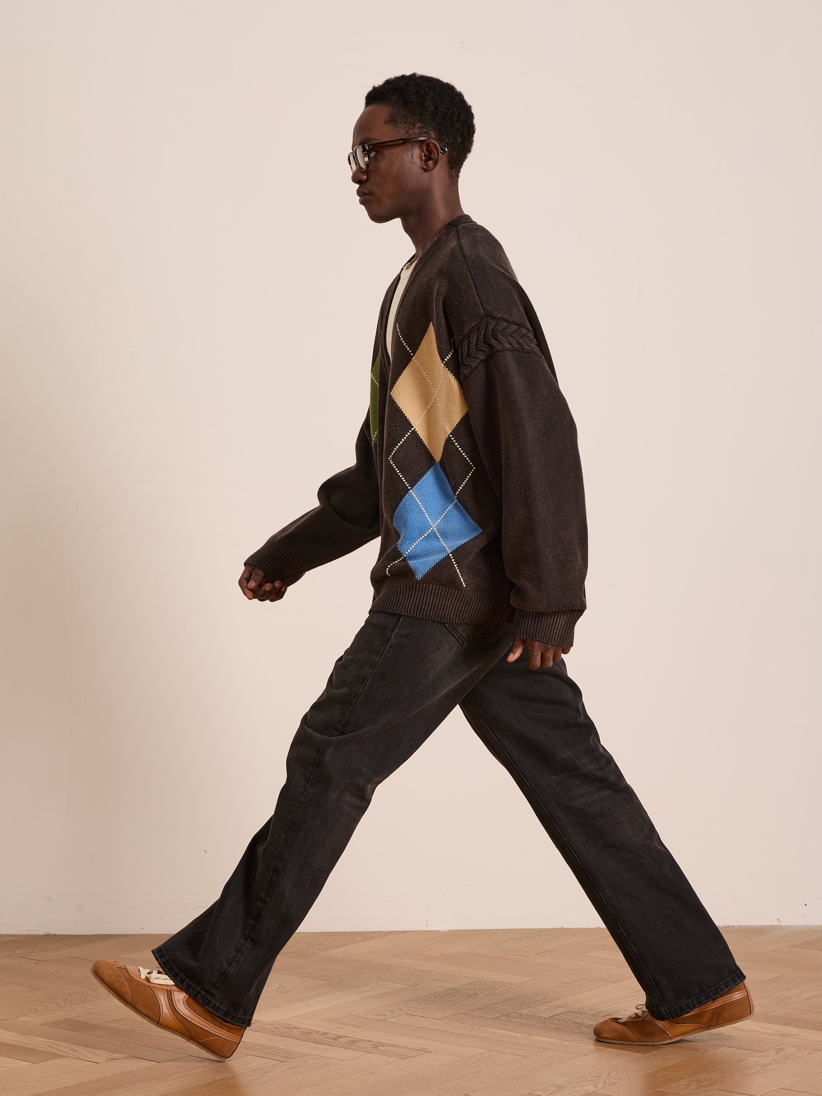 A person wearing glasses, a FOUND Argyle Knit Cardigan, dark jeans, and brown shoes walks indoors on a wooden floor against a plain background.