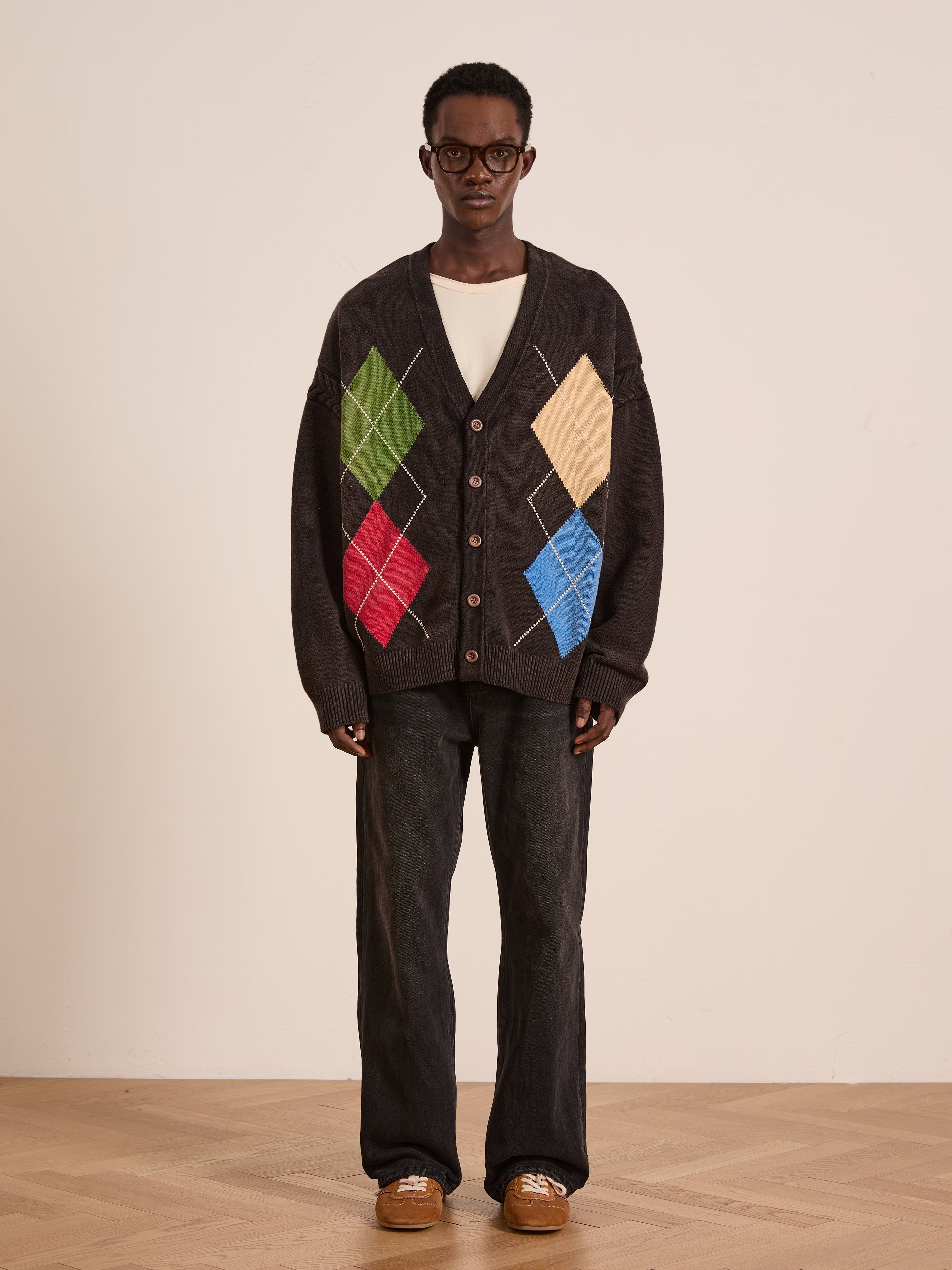 A person wears the FOUND Argyle Knit Cardigan with green, red, yellow, and blue diamonds, paired with dark jeans, brown shoes, and glasses indoors against a plain wall and wooden floor.