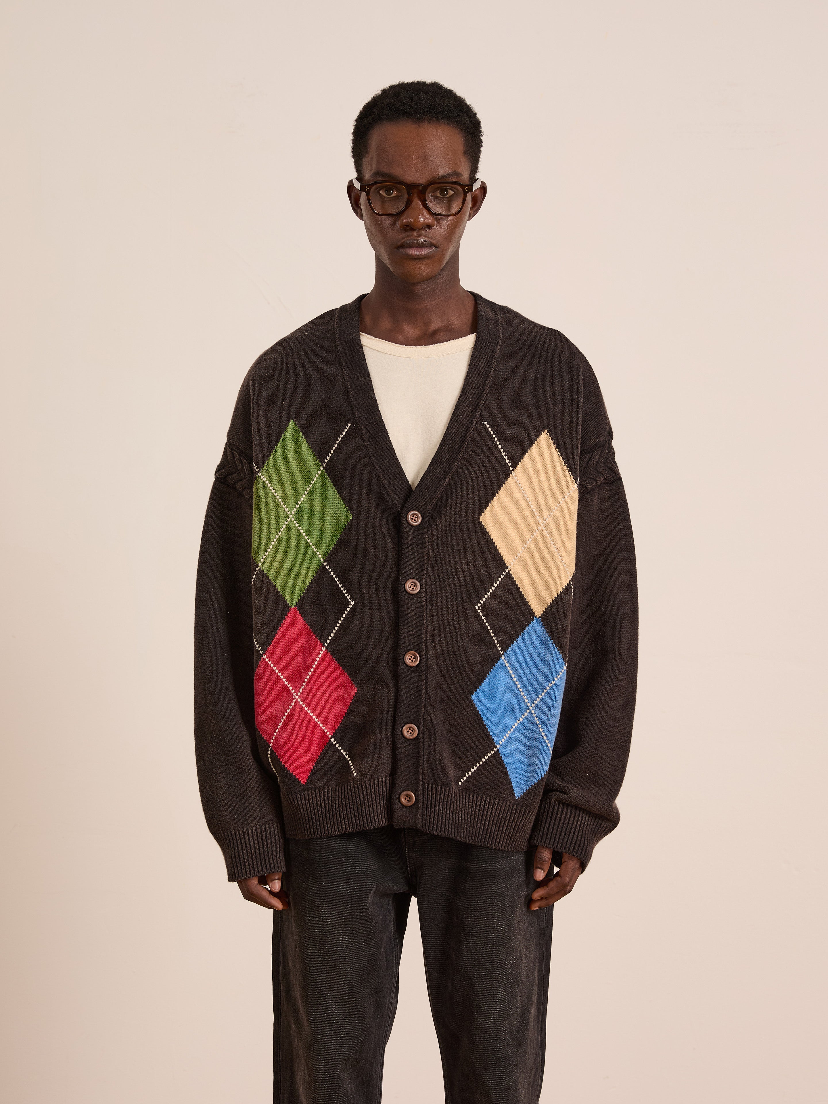 A man in glasses wears the FOUND Argyle Knit Cardigan—an oversized black sweater with green, red, yellow, and blue patterns—standing against a plain light background.