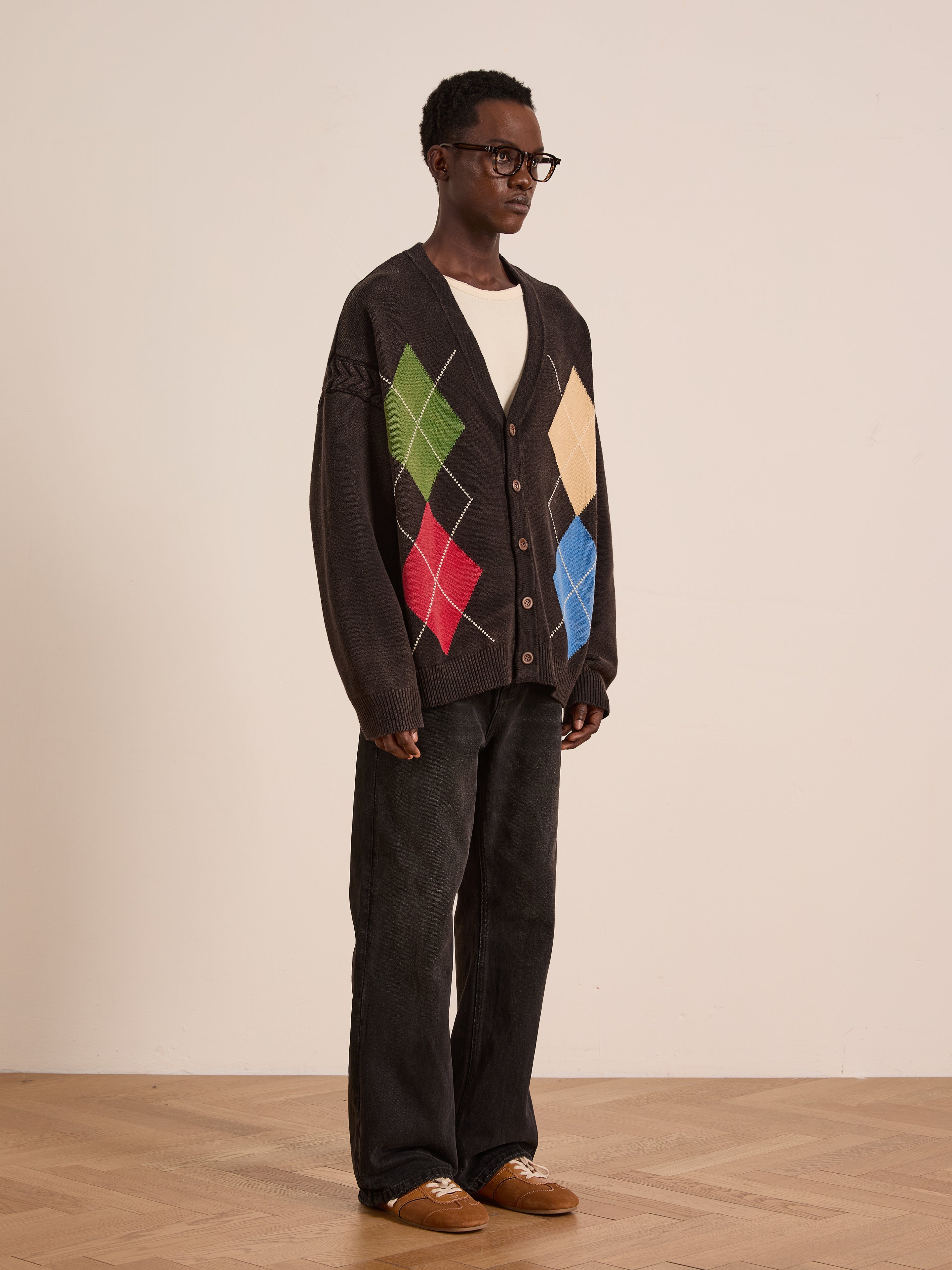 A person stands indoors wearing the FOUND Argyle Knit Cardigan, featuring colorful diamond patterns, paired with glasses, a white shirt, dark jeans, and brown shoes on a wooden floor against a plain background.