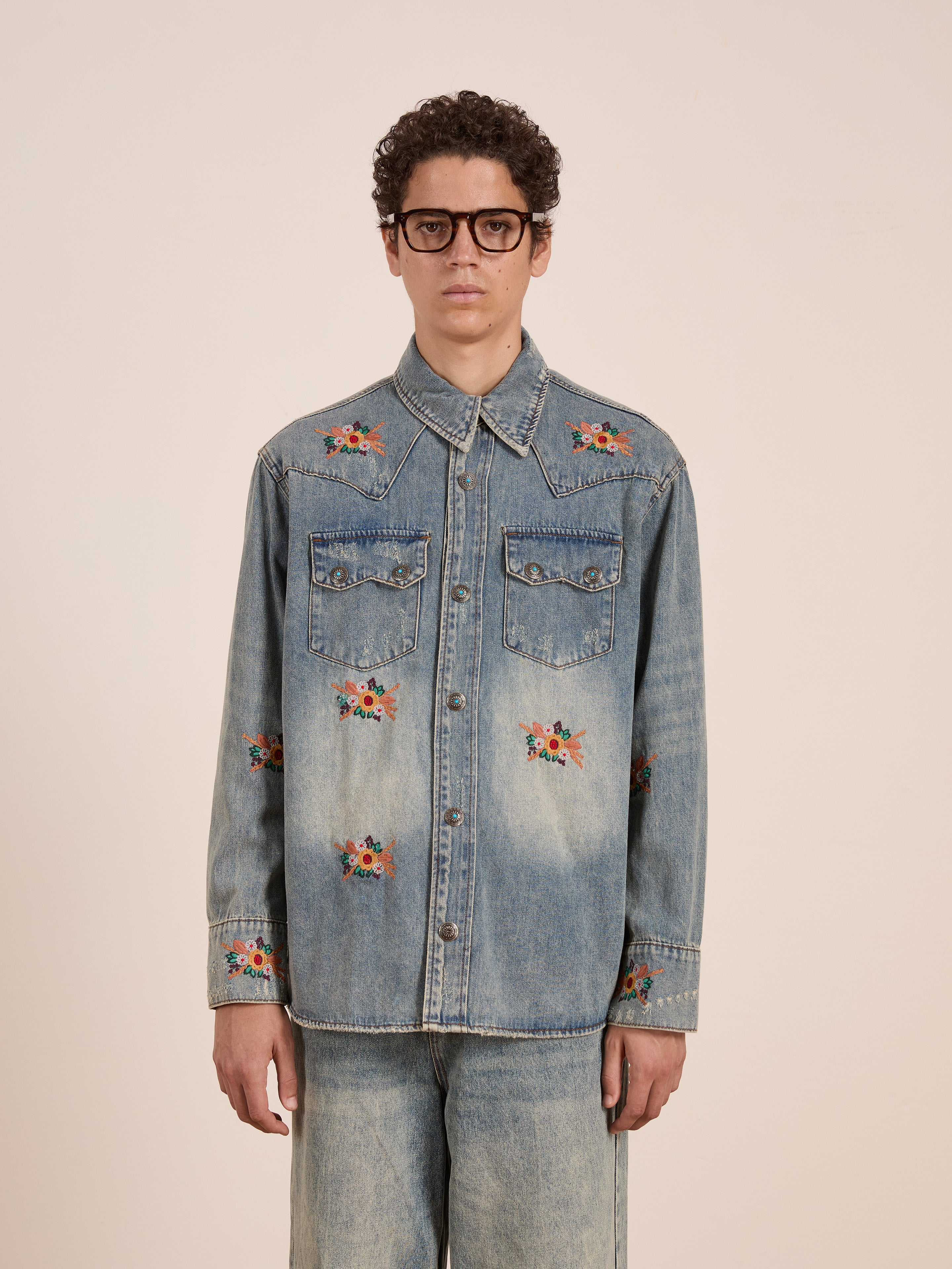 A person in the FOUND Bouquet Floral Embroidered Western Denim Shirt, matching denim pants, and large black eyeglasses stands against a plain background.