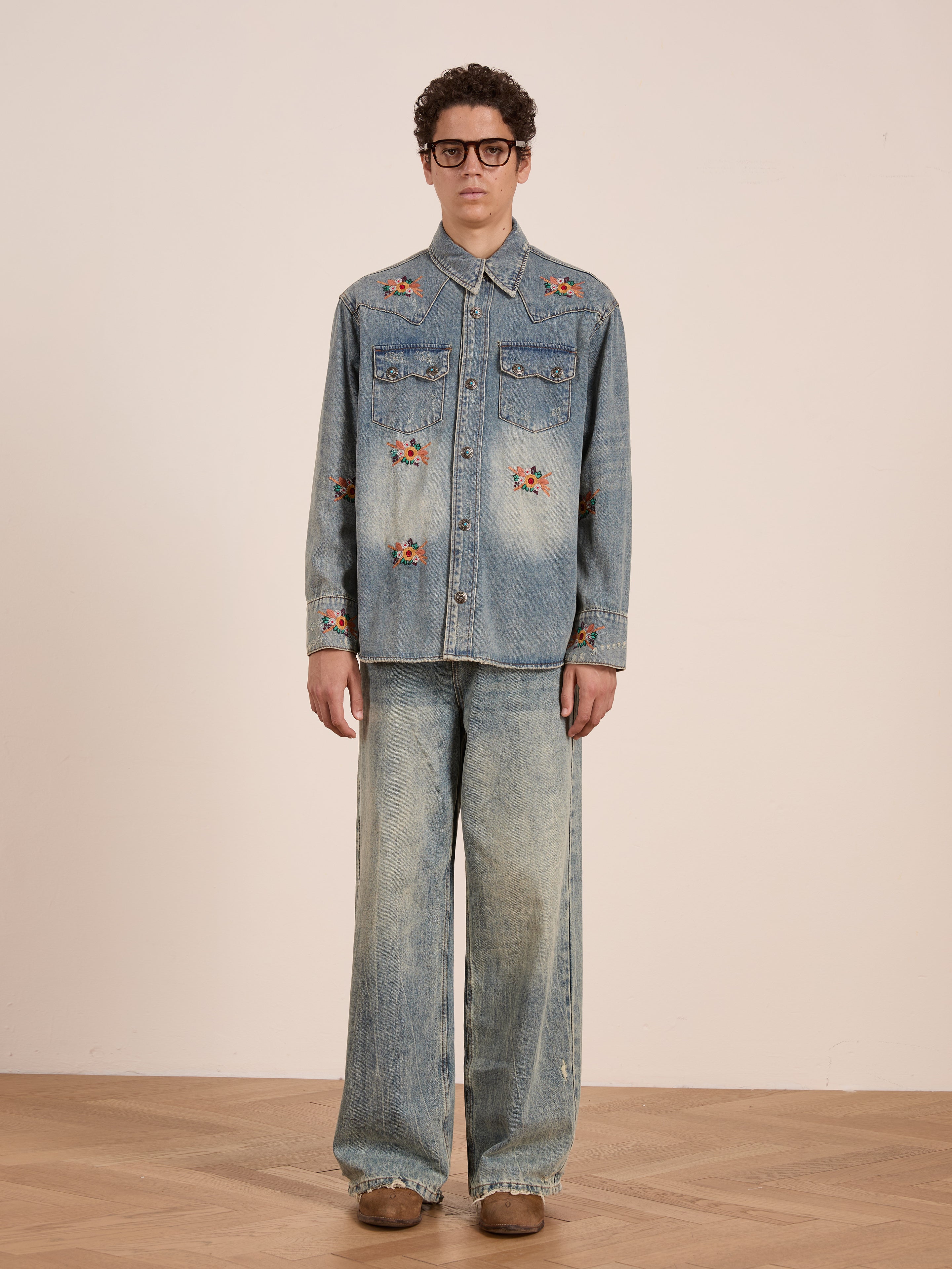 Someone stands indoors wearing the FOUND Bouquet Floral Embroidered Western Denim Shirt, matching wide-leg jeans, brown shoes, and large black glasses.