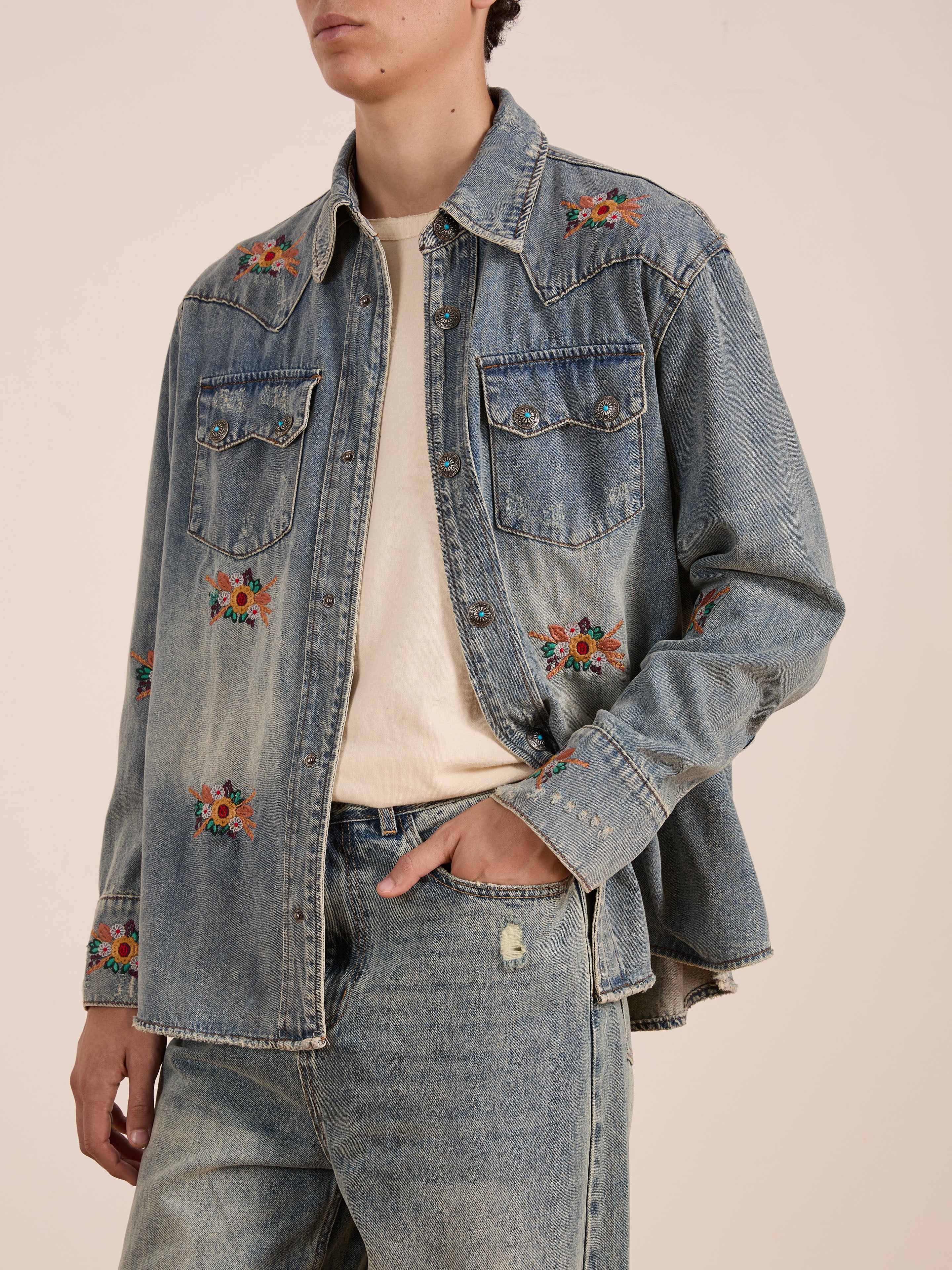 A person wears the FOUND Bouquet Floral Embroidered Western Denim Shirt over a beige t-shirt and matching relaxed fit denim jeans, with one hand in their pocket.