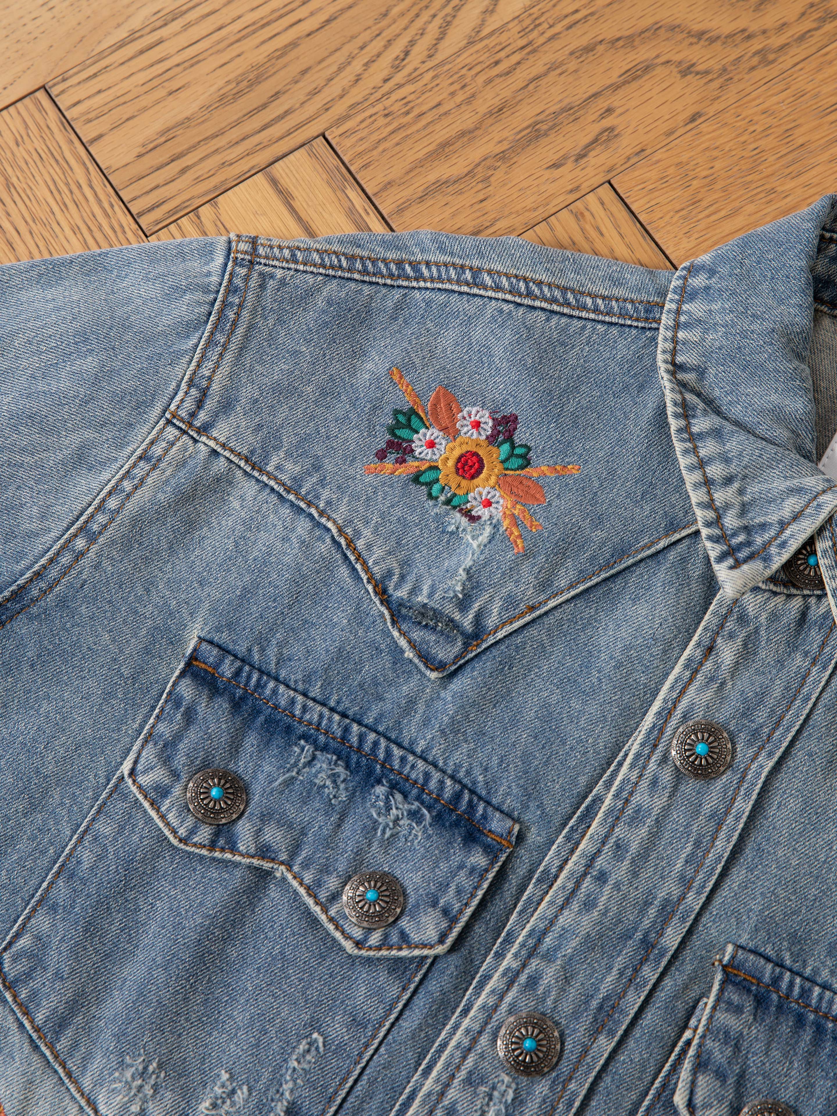 A close-up of the FOUND Bouquet Floral Embroidered Western Denim Shirt, featuring floral embroidery on the chest, metal buttons, and distressed details, laid out on a wooden floor.