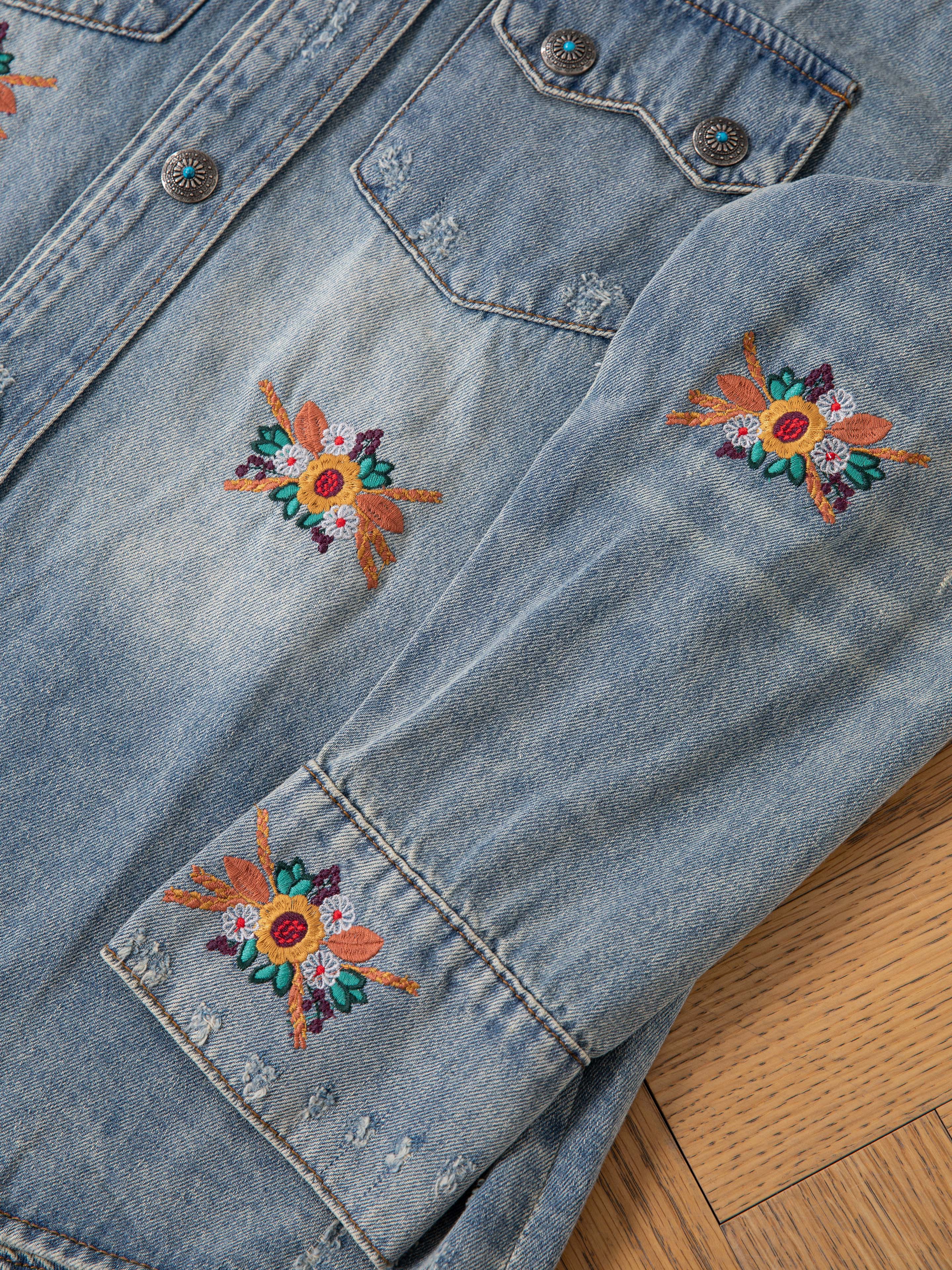 Close-up of the FOUND Bouquet Floral Embroidered Western Denim Shirt in light blue, featuring floral embroidery on the sleeve and chest. Crafted in a relaxed fit and displayed flat on a wooden surface.