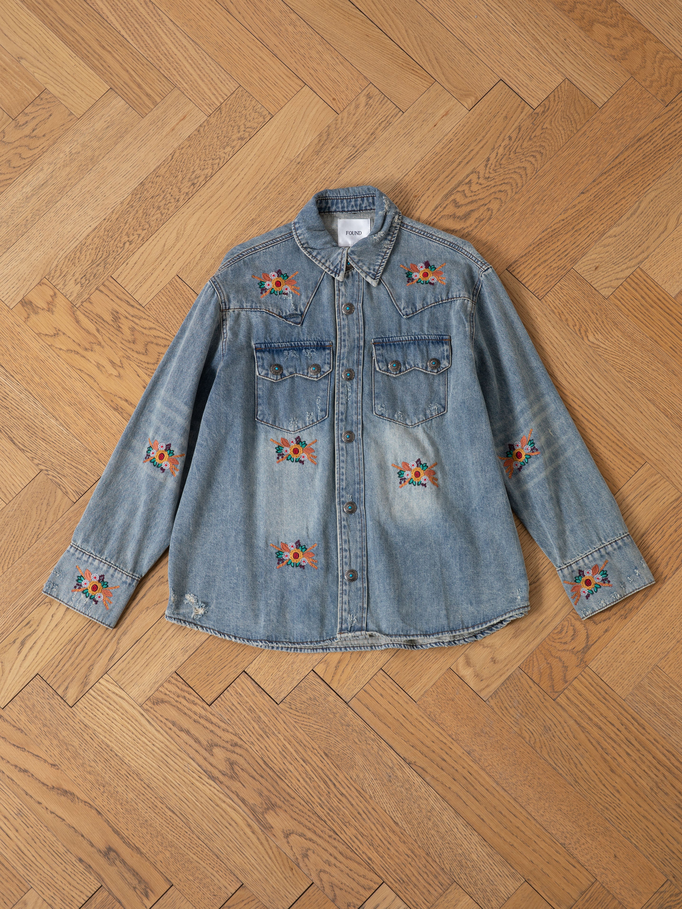 The FOUND Bouquet Floral Embroidered Western Denim Shirt features light blue denim with floral embroidery on the chest, sleeves, and lower front in a relaxed fit, shown displayed on a wooden herringbone floor.