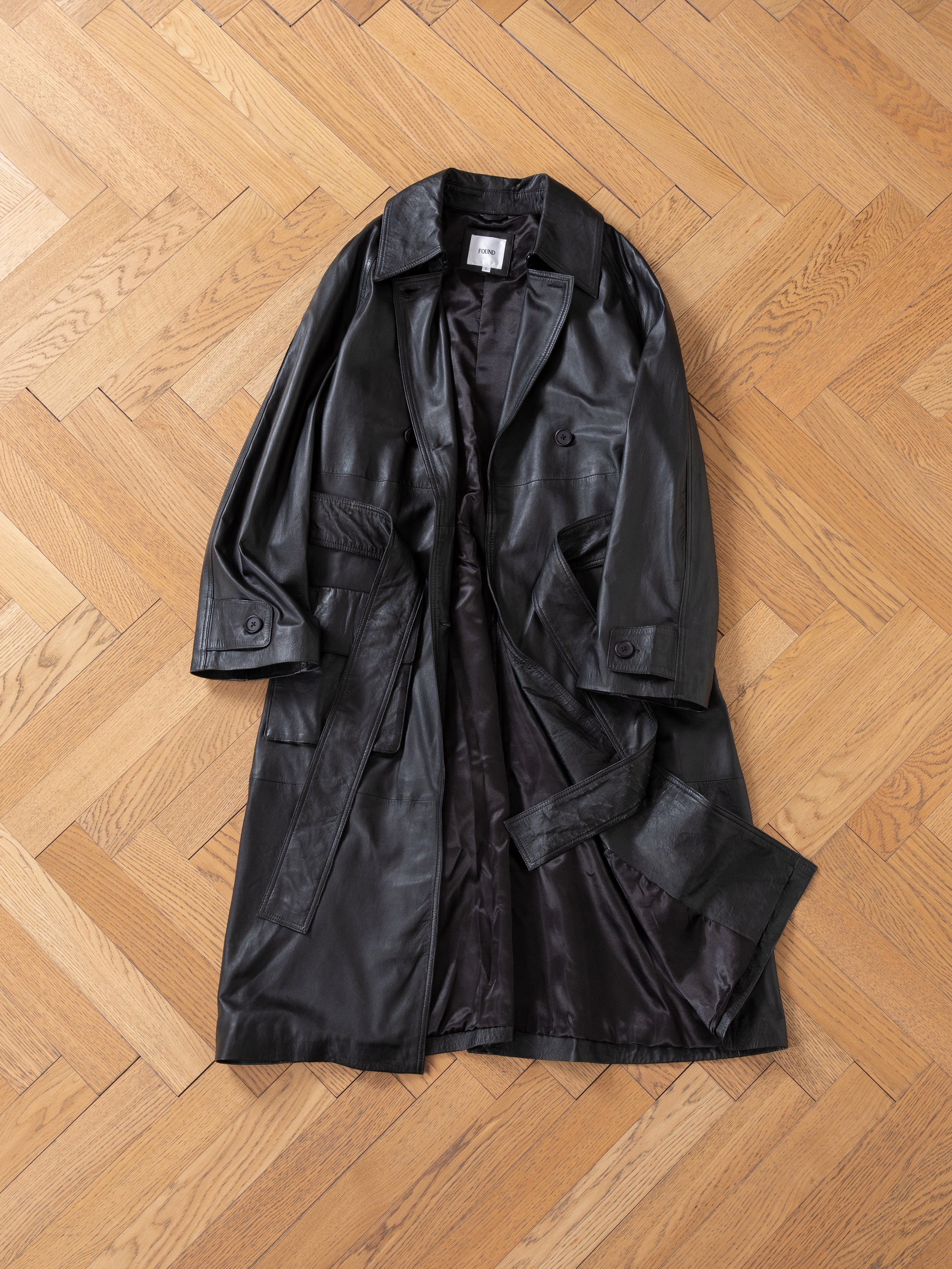 The FOUND Leather Trench Coat, crafted from premium cowhide and featuring a belt, is laid flat on a light wood herringbone floor.
