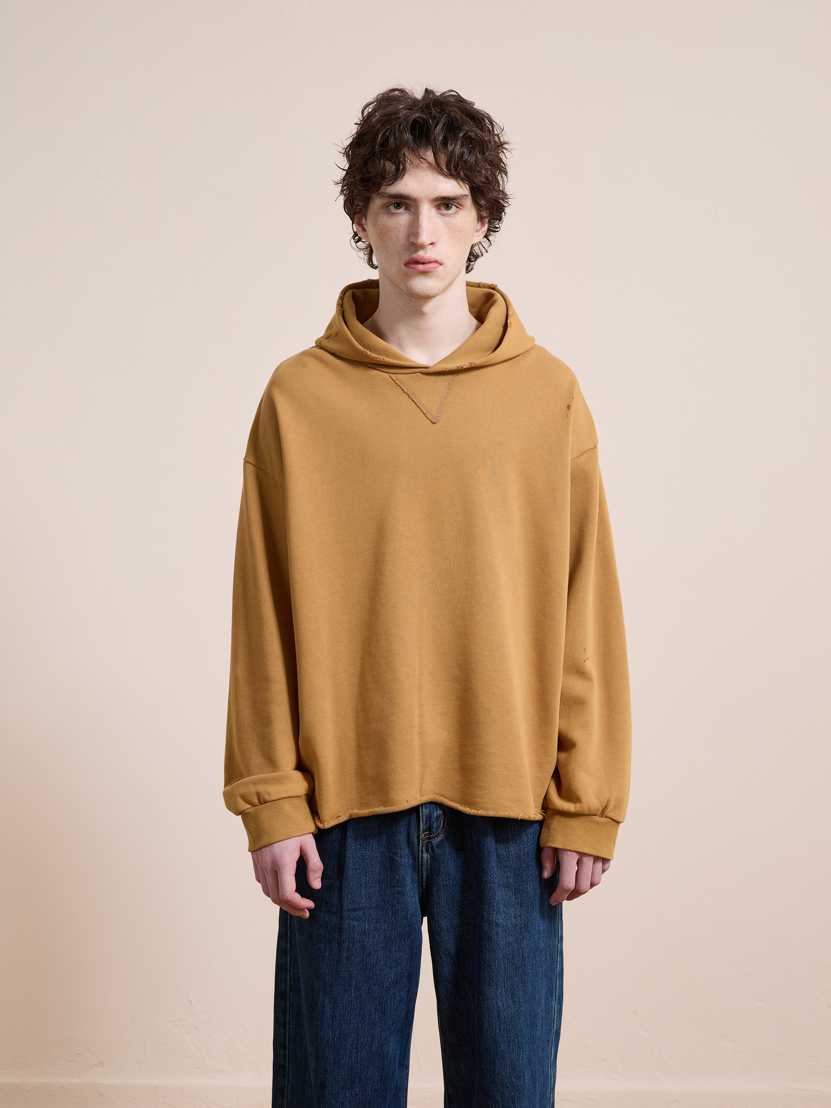 A person with curly brown hair wears the FOUND Beadwork Logo Hoodie in mustard yellow and blue jeans, standing against a plain beige background.