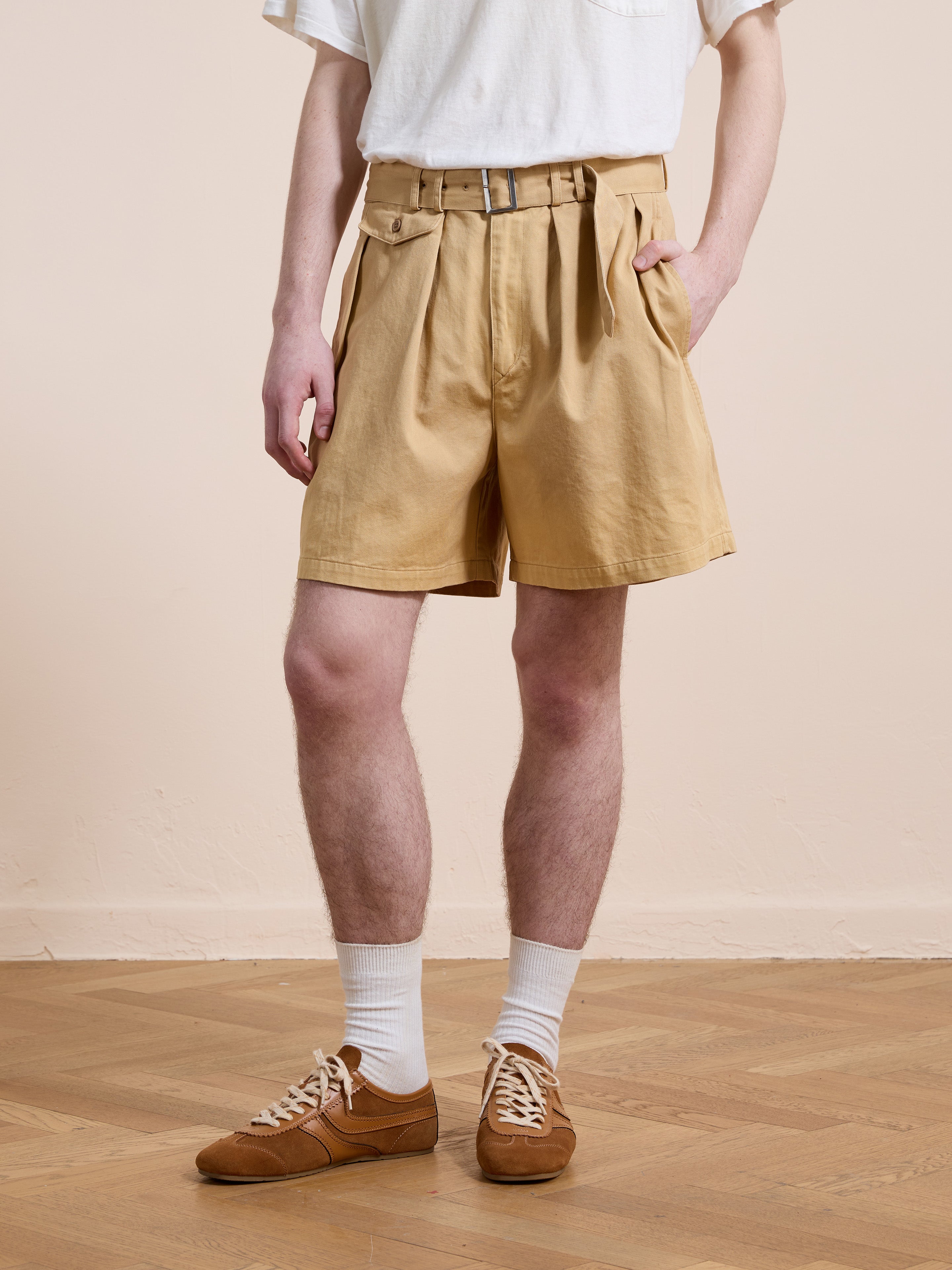 A person wearing a white t-shirt, brown Found Belted Twill Pleated Shorts, white socks, and brown sneakers stands on a wooden floor against a light beige wall.