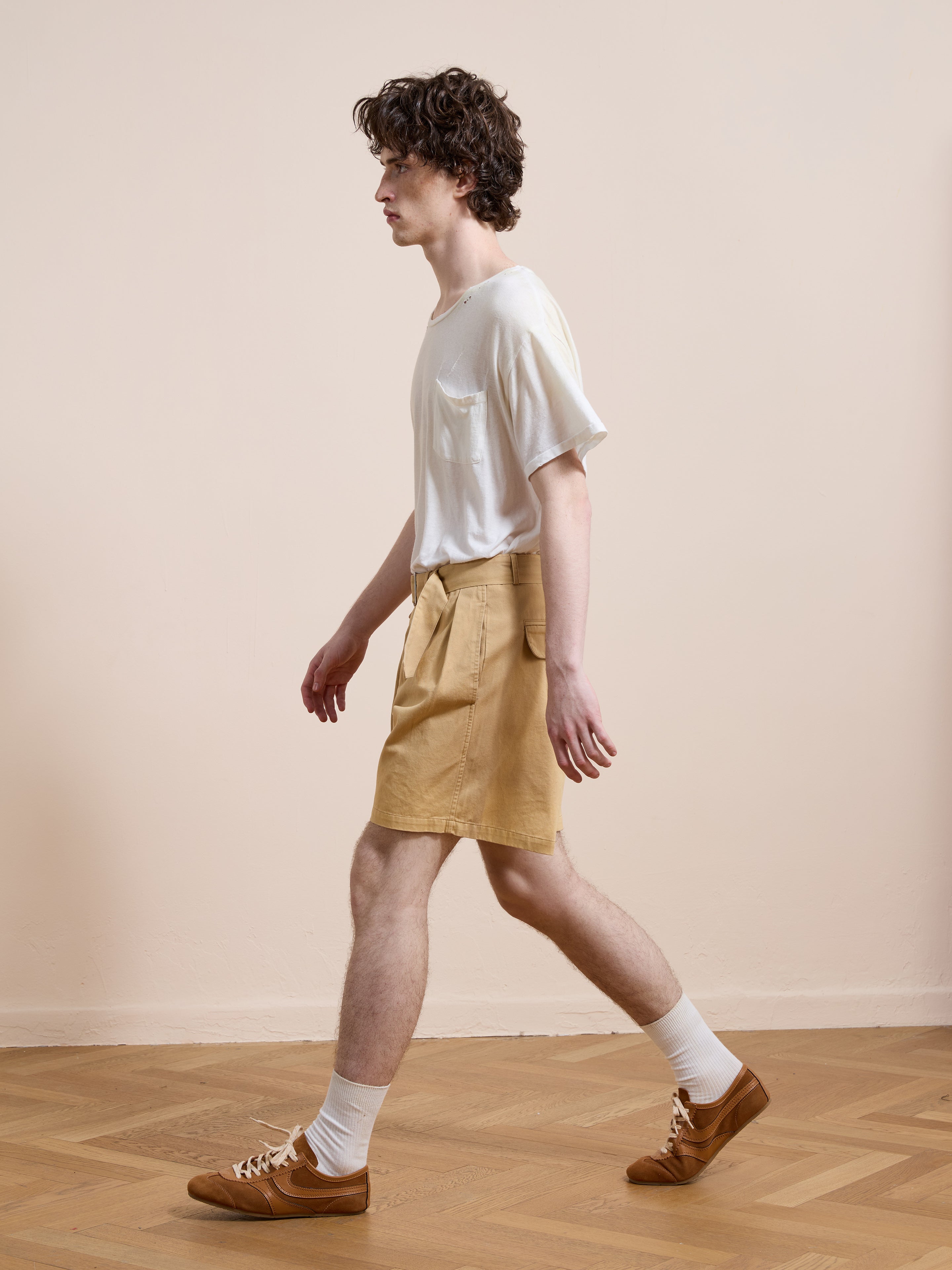 A young man with curly hair walks indoors on a wooden floor against a beige backdrop, wearing a white T-shirt, white socks, brown shoes, and Found Belted Twill Pleated Shorts.