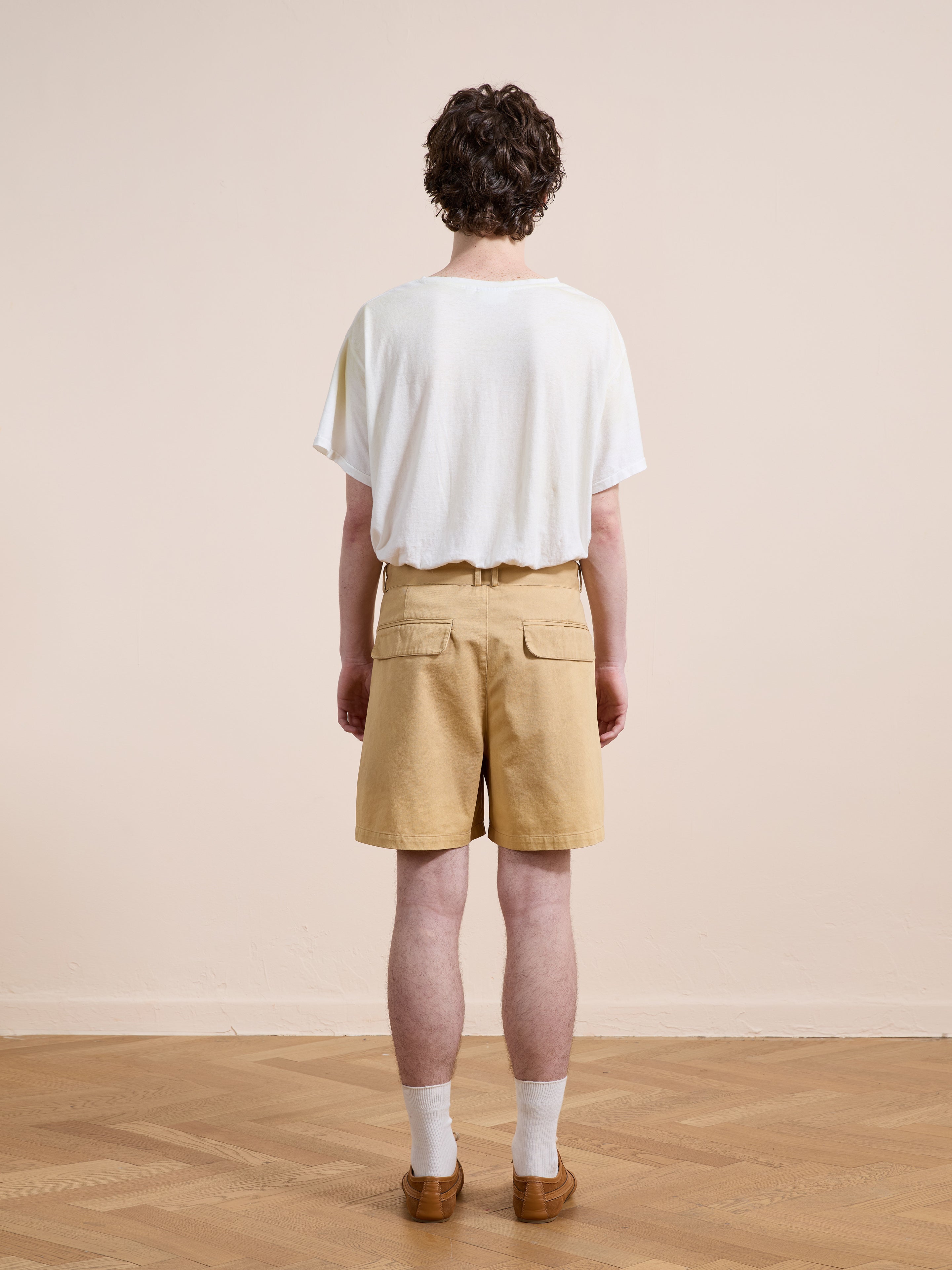 A person with short curly hair faces away, wearing a loose white T-shirt, Found's Belted Twill Pleated Shorts, white socks, and brown shoes on a wooden floor with a plain beige background.