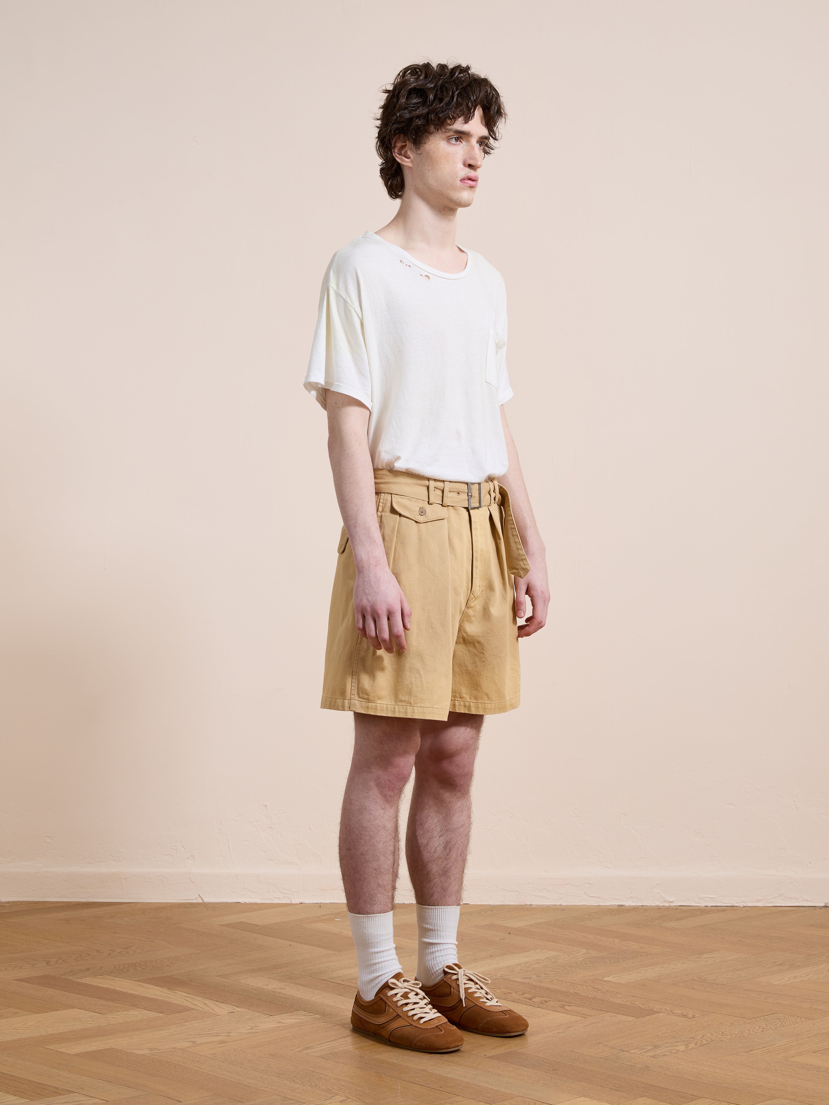 A person stands indoors against a plain beige background wearing Found's Belted Twill Pleated Shorts, a white t-shirt, white socks, and brown sneakers, facing slightly to the side.