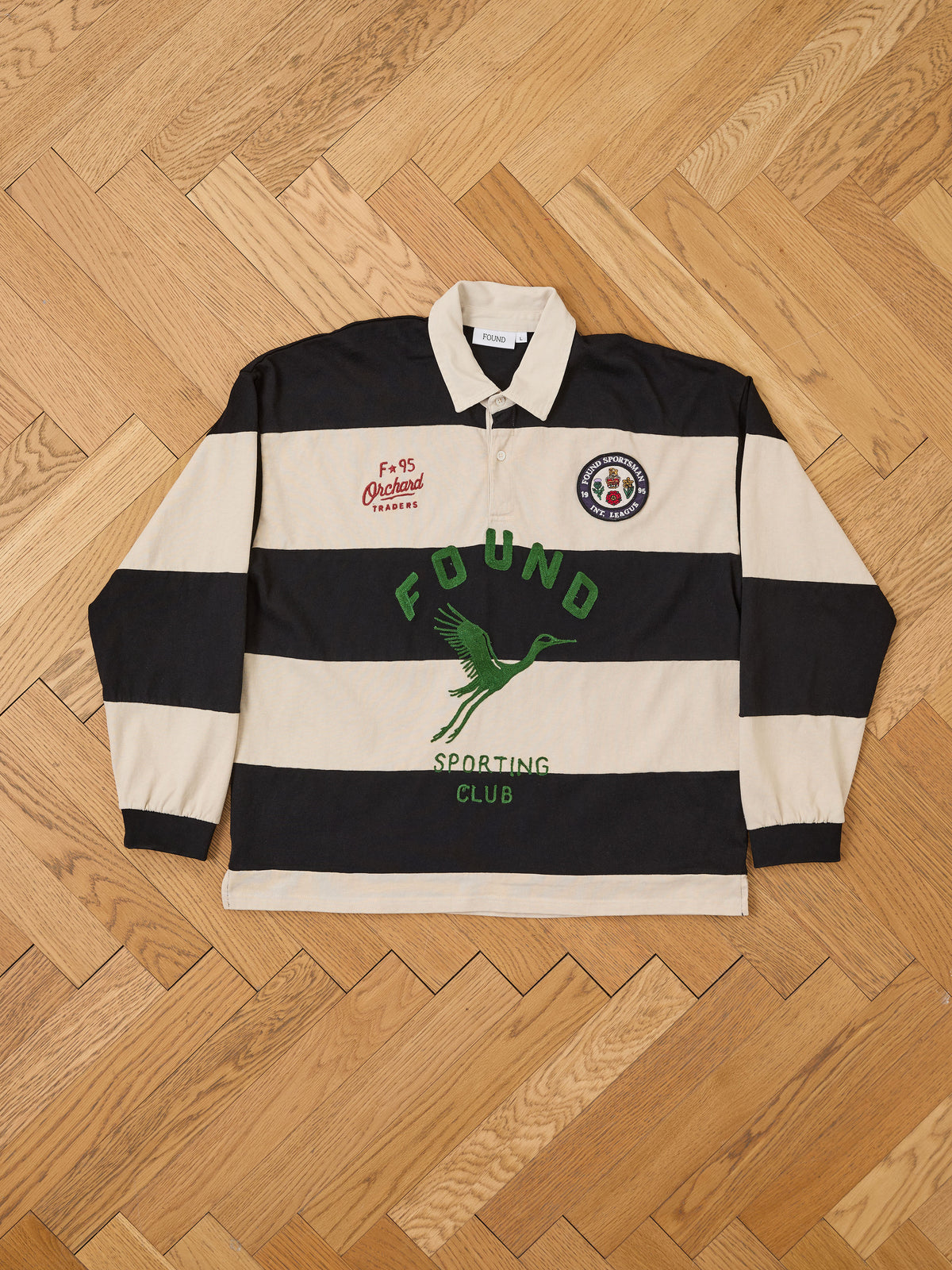 The FOUND Bird Logo Rugby is a long-sleeve unisex cotton top with black and beige stripes, green text, a bird graphic, and chest patches.