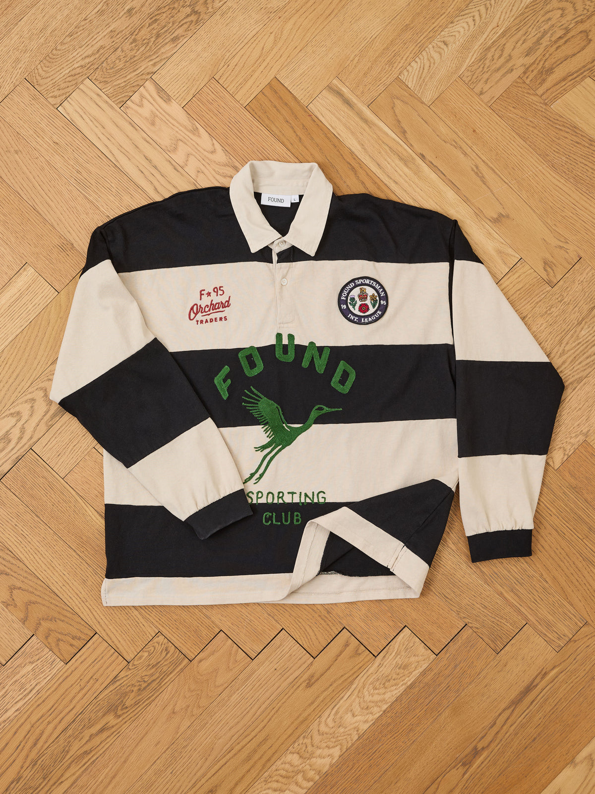 Bird Logo Rugby by FOUND: long-sleeve unisex relaxed fit shirt with black/beige stripes, green branding, and embroidered patches laid flat on wood floor.