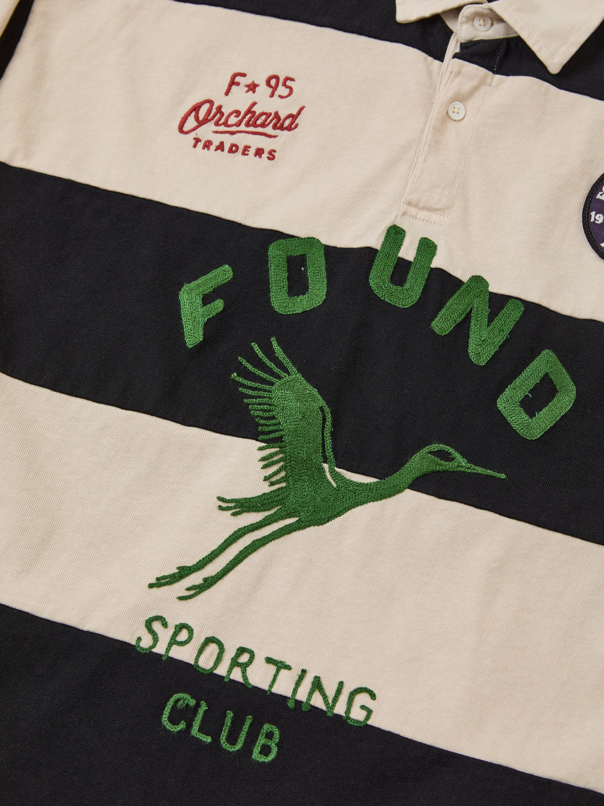 The Bird Logo Rugby by FOUND is a unisex black and beige striped shirt with “FOUND”, a green crane, and “F*95 Orchard TRADERS” embroidery above the stripes.