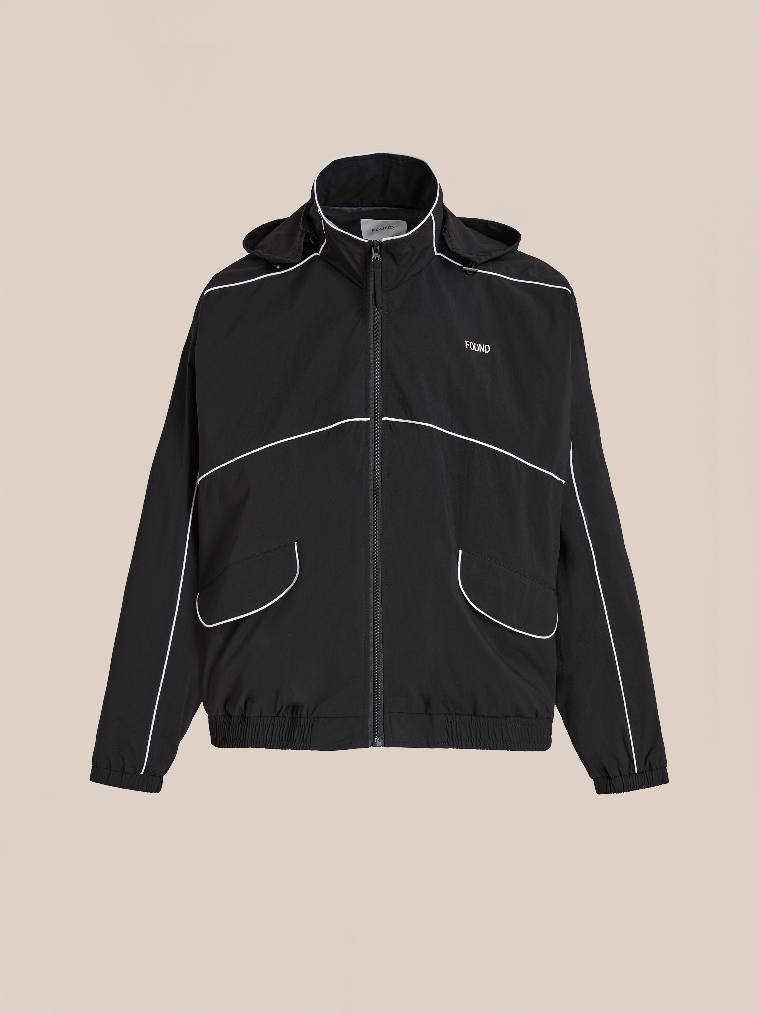 The FOUND Arena Stripe Track Jacket is a unisex black zip-up made from lightweight technical fabric, featuring white piping, two front pockets, elastic cuffs and hem, and a small chest logo. Shown on a beige background.