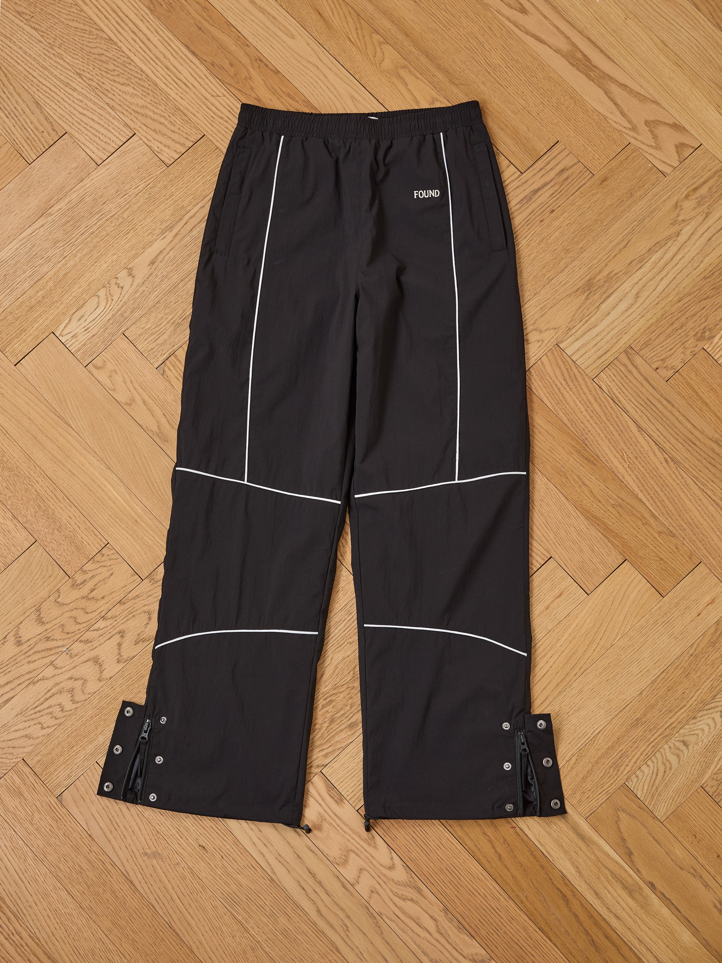 Found Arena Stripe Track Pants in black with white piping, adjustable waistband, snap-button cuffs, and "FOUND" text on the front, displayed flat on a wooden herringbone floor.